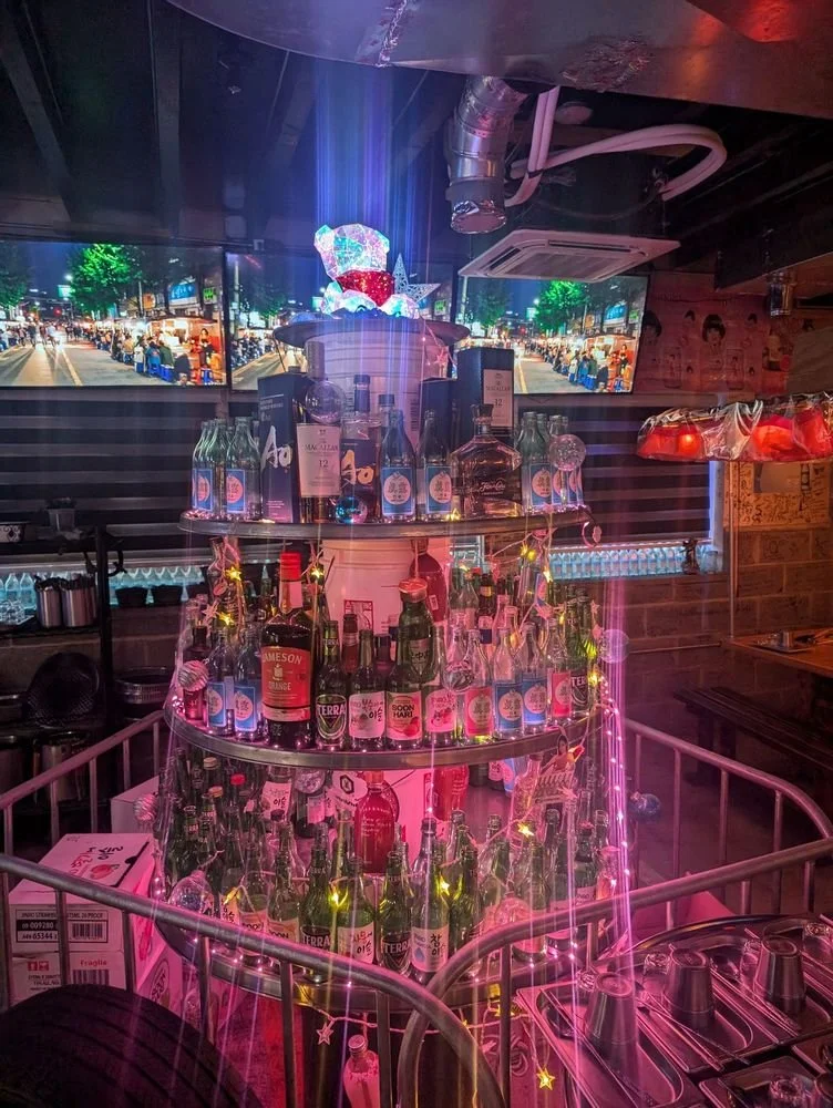 Bar display with multiple bottles of alcohol arranged in a circular tiered setup, illuminated with colorful lights and star-shaped decorations, with a city street scene on multiple TV screens in the background.