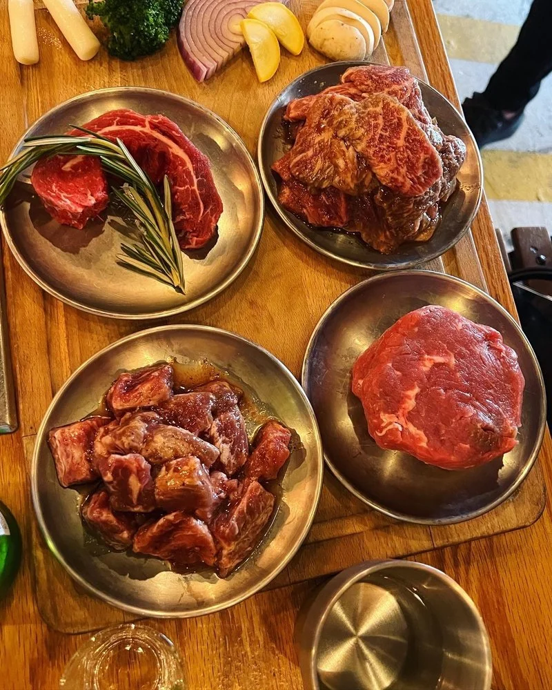 Assorted raw meats, vegetables, and seasonings on a wooden table, including steaks, beef chunks, a large beef roast, onions, garlic, herbs, and vegetables.