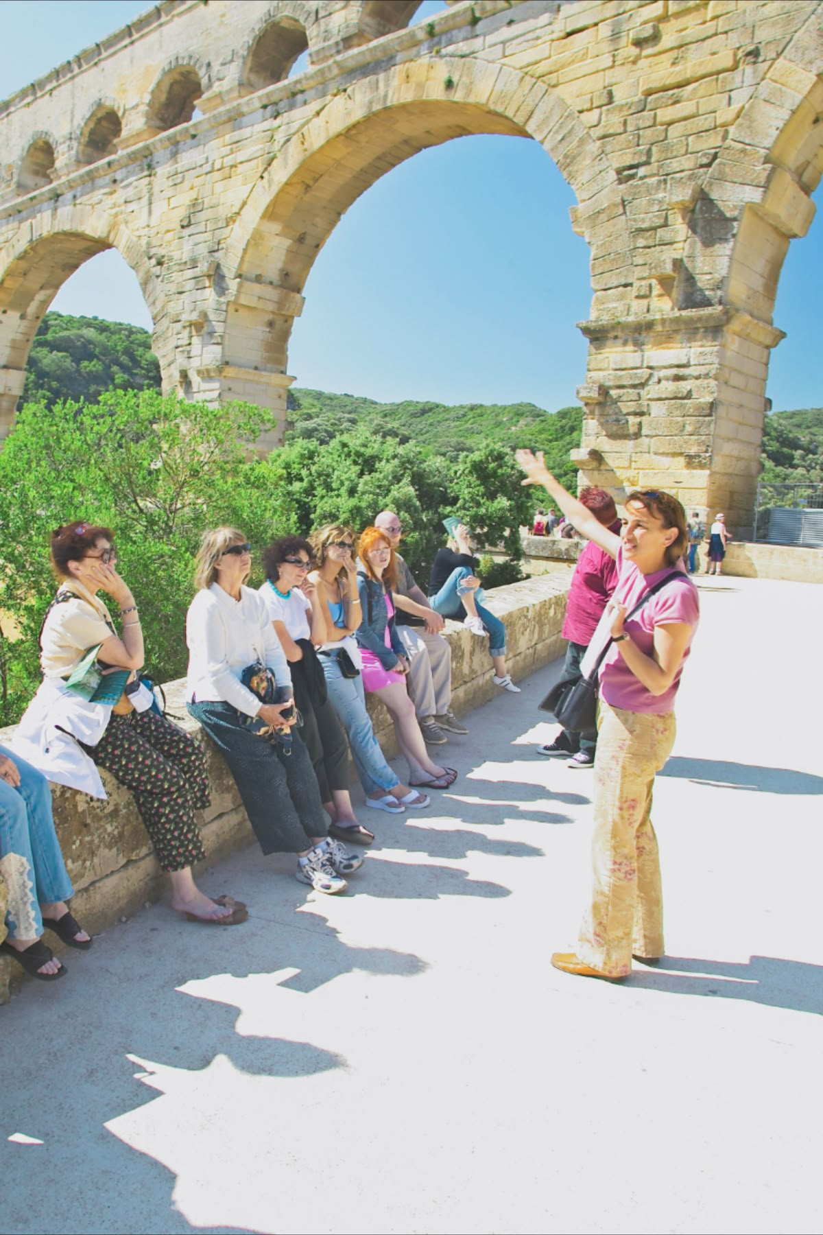 A tour guide speaking to a group of tourists sitting on a stone ledge under a large ancient stone Roman aqueduct arch, with green hills and trees in the background on a sunny day.