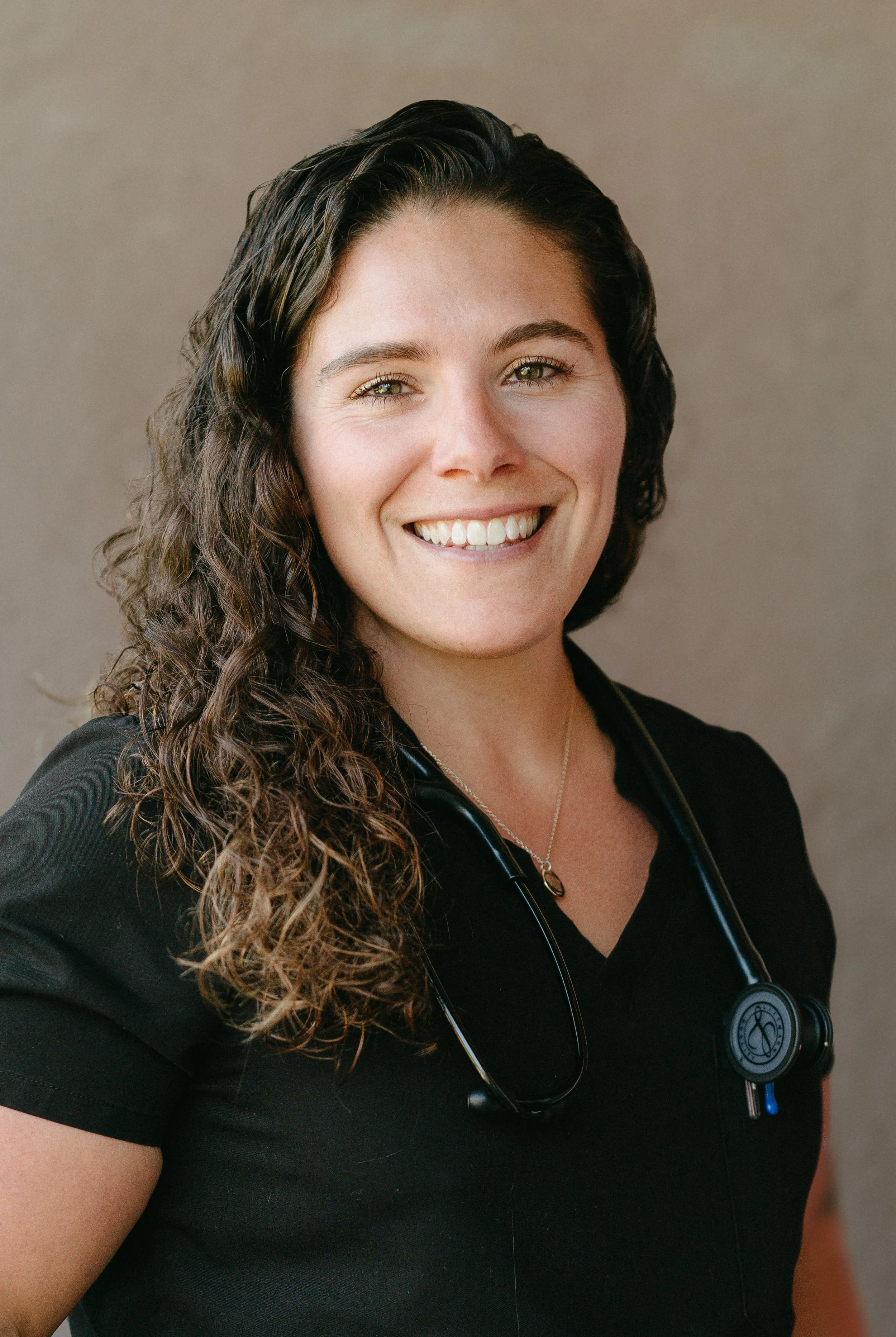 A smiling woman with curly brown hair wearing a black shirt and a stethoscope around her neck.