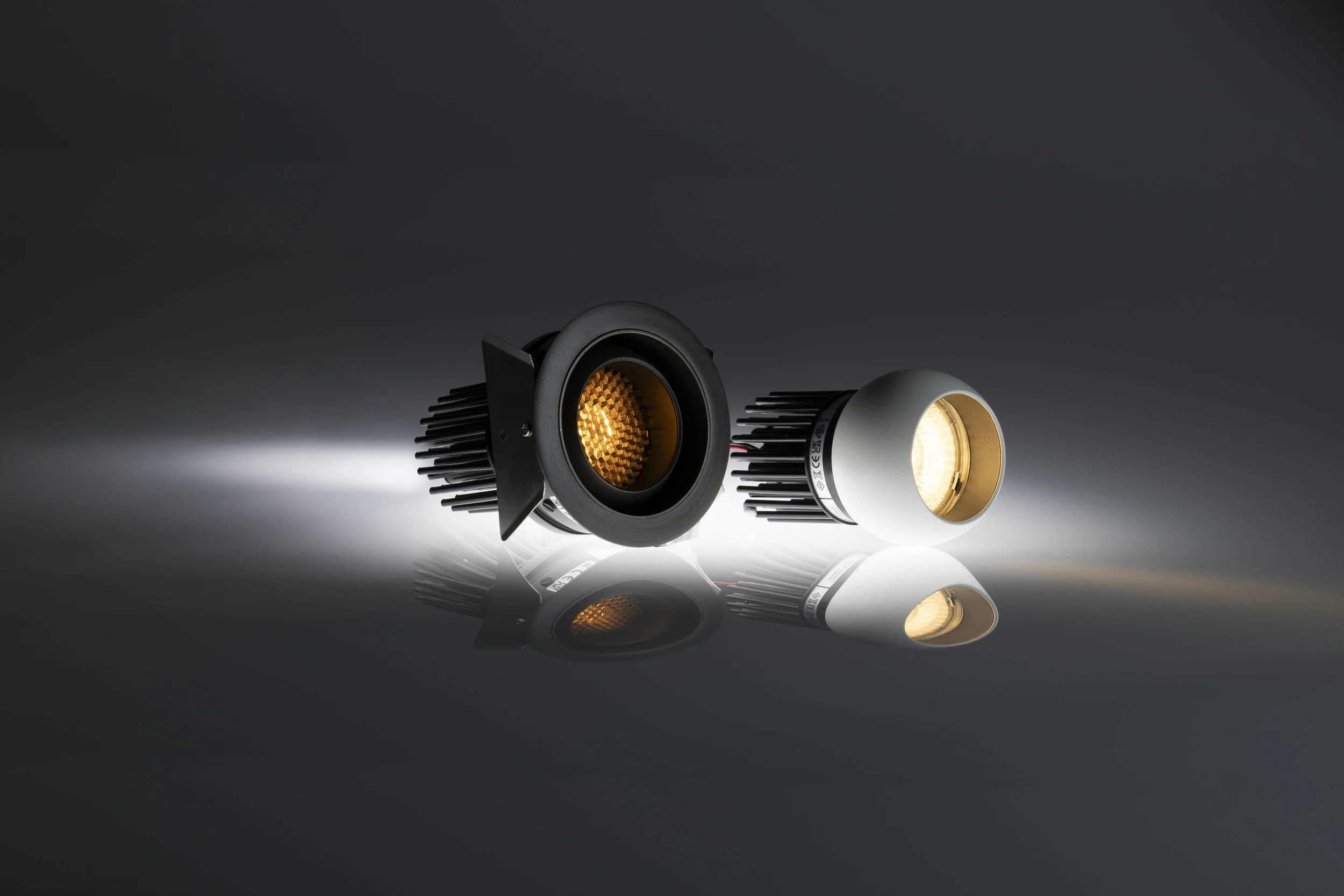 Two LED light fixtures with black and white housing, lying on a reflective surface against a dark background, emitting light.
