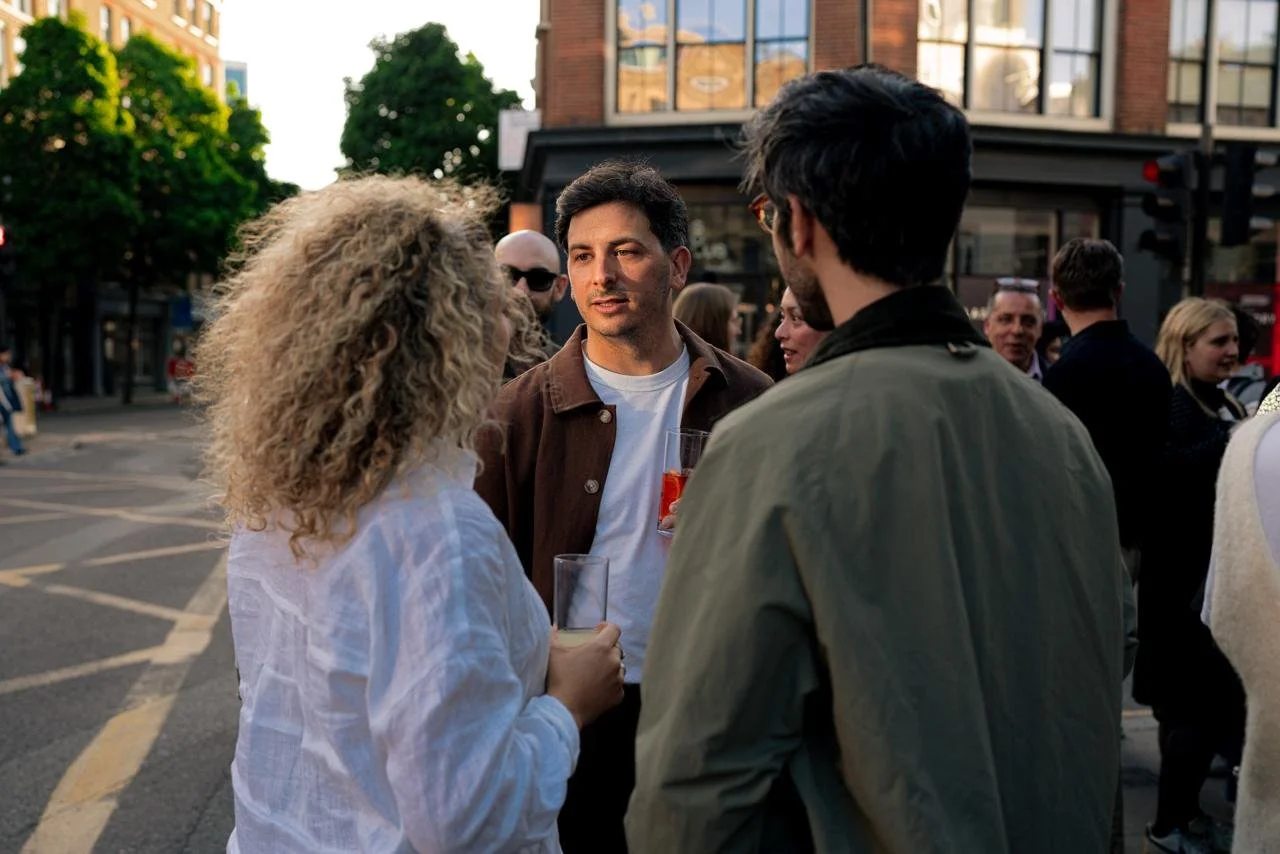 A group of people socializing outdoors in an urban area during the daytime. A woman with curly blonde hair and a man with dark hair and glasses are engaged in conversation, holding drinks. Other individuals are mingling in the background.