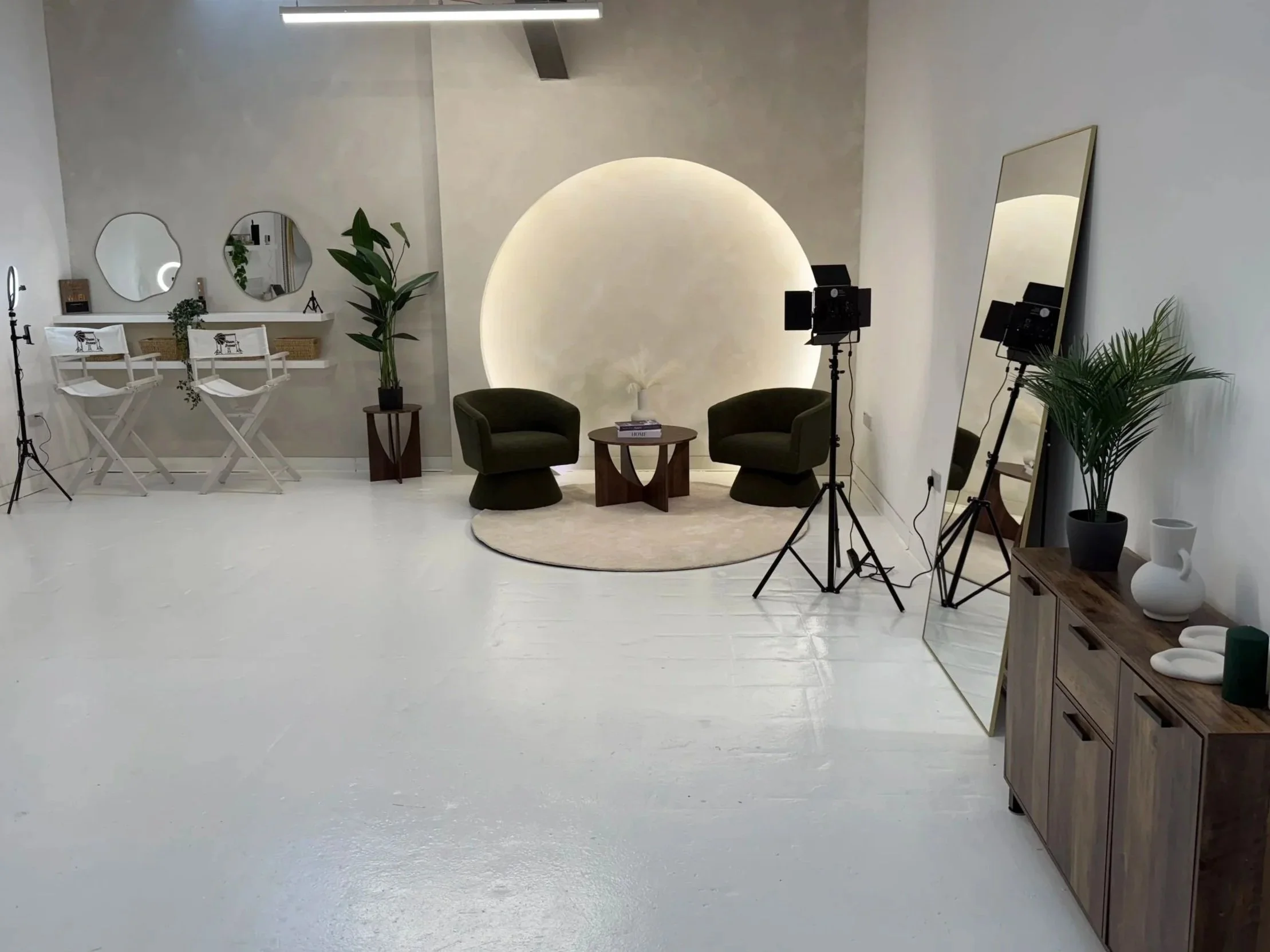 A photography studio with two green armchairs and a small round table with books, set against a circular illuminated wall. Studio lighting equipment and mirrors are visible, along with plants and decorative items on a wooden sideboard.