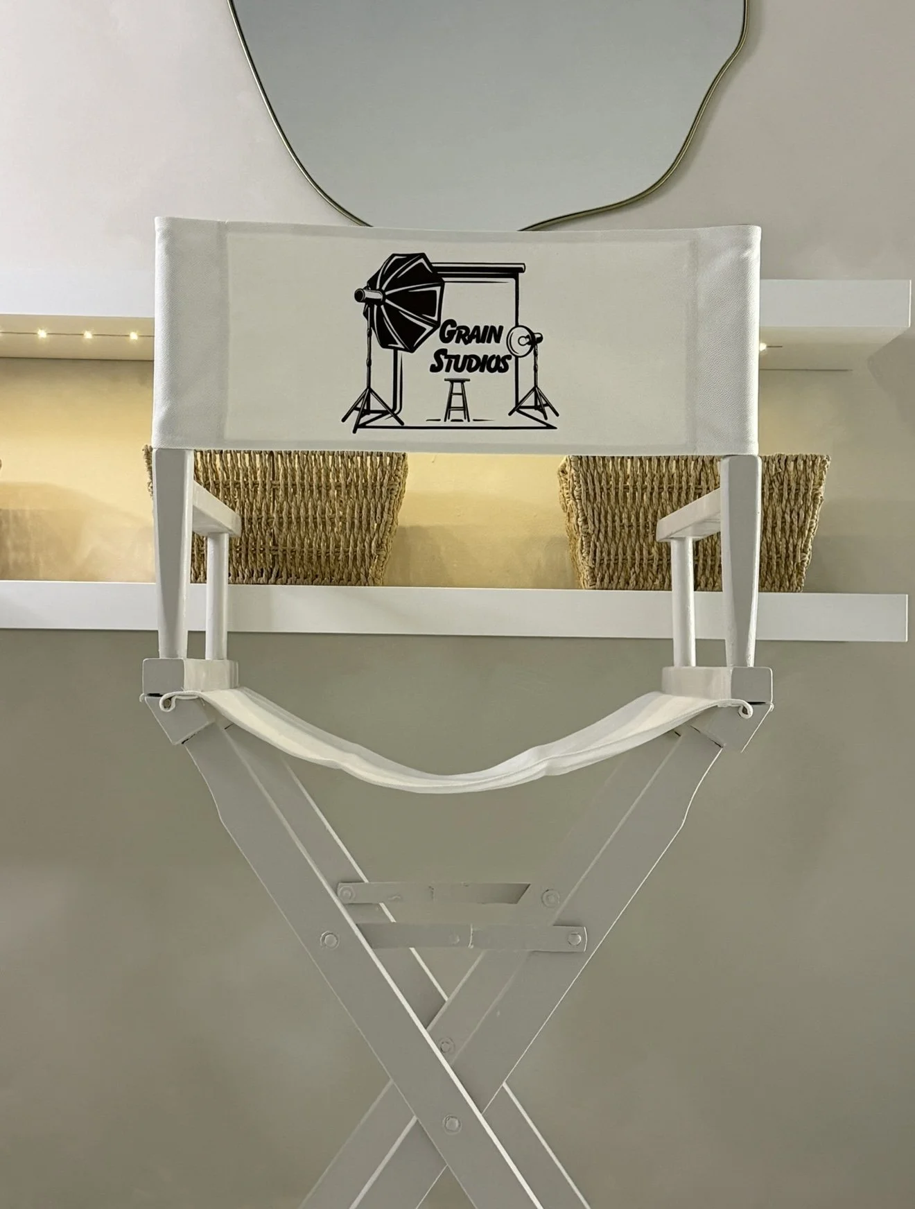 Director's chair with 'GRAIN STUDIOS' logo, a photography setup with umbrella light and backdrop, hanging mirror, wicker baskets, and a white shelf.