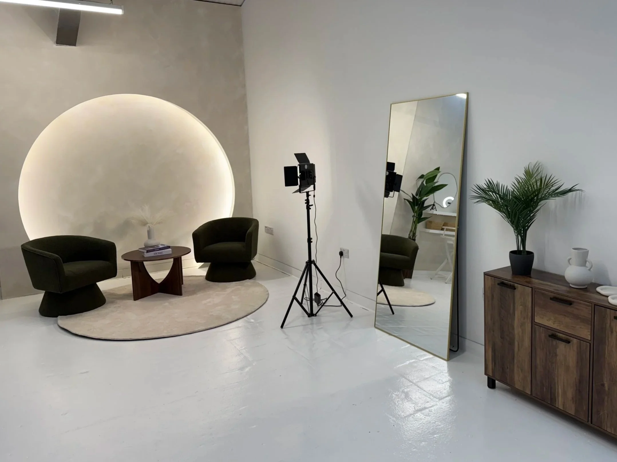 Minimalist photography studio with two black chairs, a small wooden table, a large mirror leaning against the wall, a studio light with a stand, and decorative plants and vases.