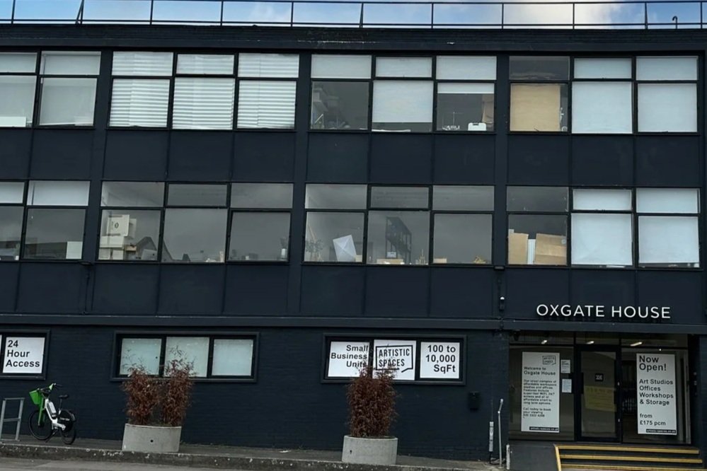 Front of OXGate House building with large windows, signs for art studios and workshops, and a bicycle parked outside.