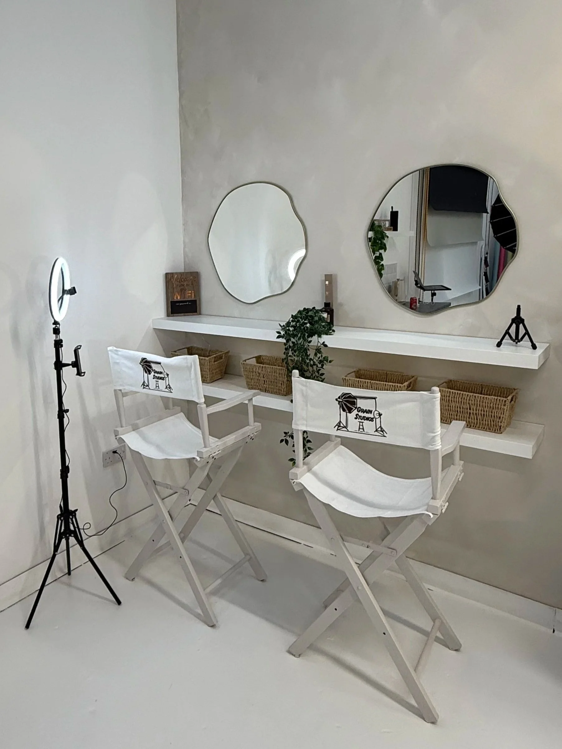 A photography studio corner with two white director chairs in front of a white wall, which has two abstract-shaped mirrors and a floating white shelf holding wicker baskets, a small plant, and decorative items. There is a ring light on a stand to the left of the chairs.