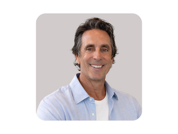 A middle-aged man with dark, curly hair and a friendly smile, wearing a light blue button-up shirt over a white t-shirt, against a plain gray background.