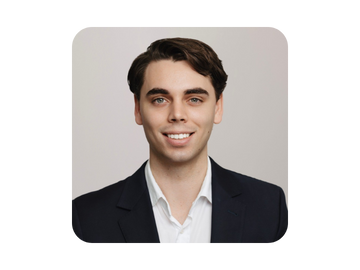 A young man in a dark suit and white shirt smiling at the camera against a plain background.