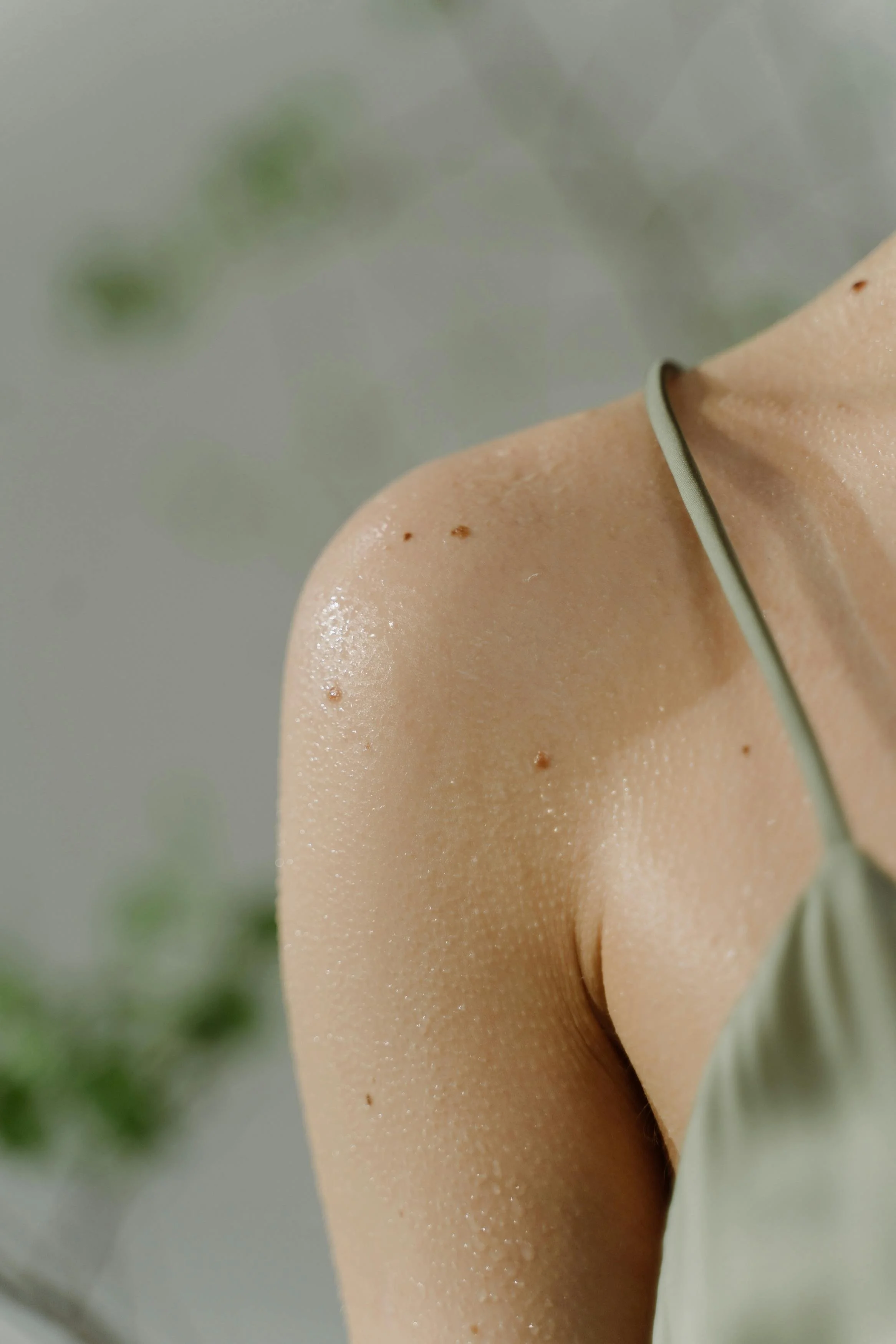 A close-up of a person's shoulder and upper arm, with small water droplets on the skin, wearing a light-colored spaghetti strap top.