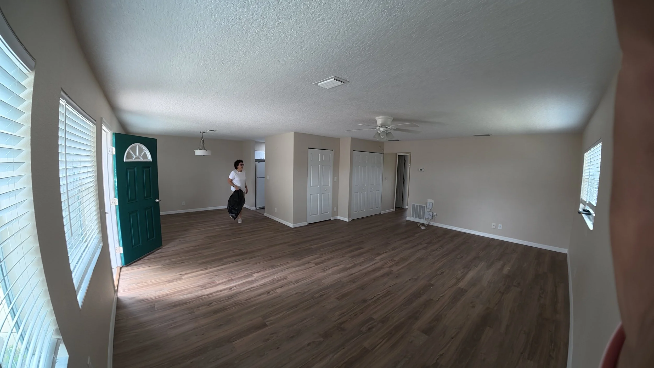 Empty living room with hardwood floors, multiple windows, a person holding a black bag, and white closet doors.