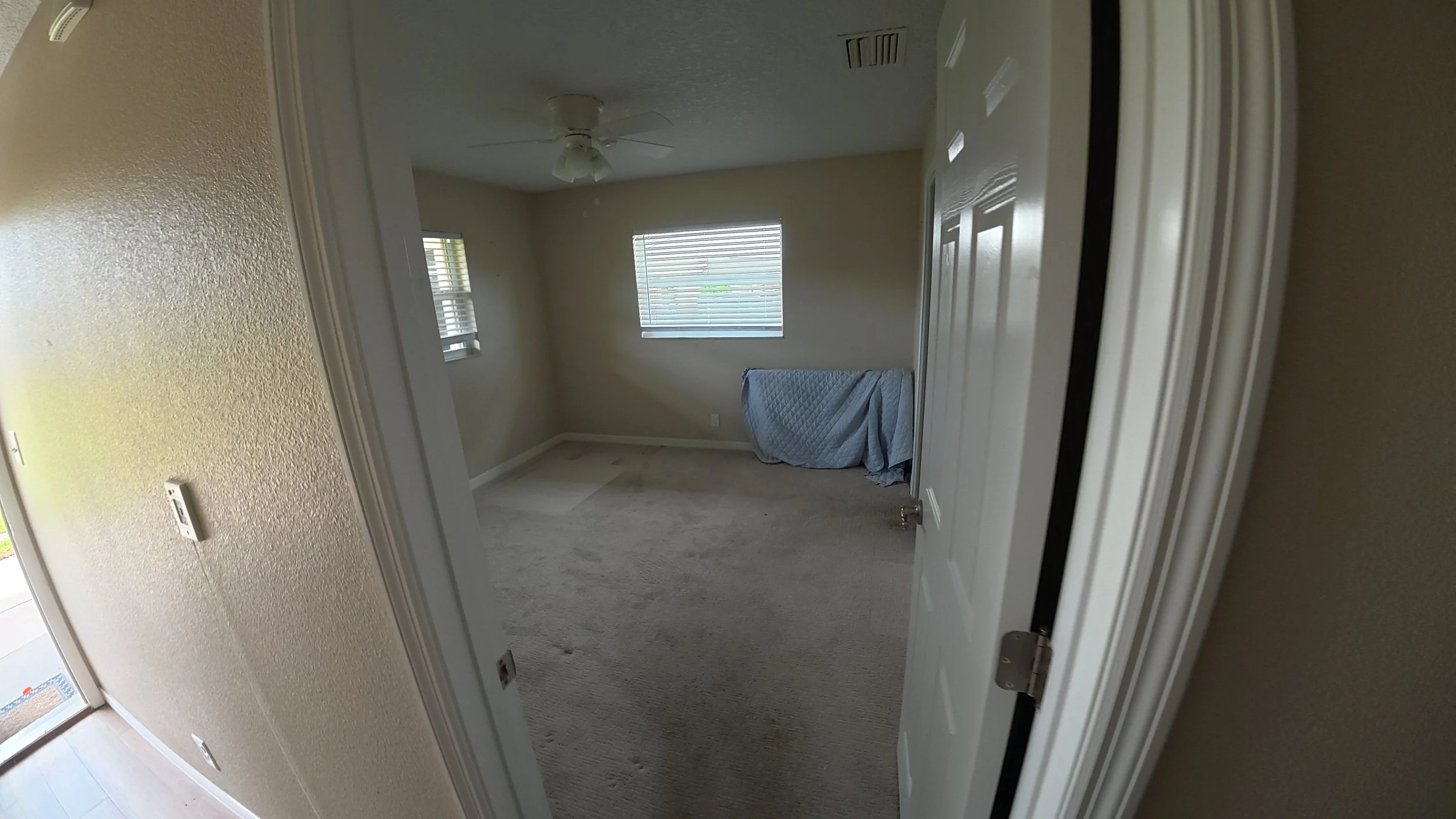 Empty bedroom with beige walls, two windows with blinds, ceiling fan, and a mattress covered with a blue quilted cover on the floor.