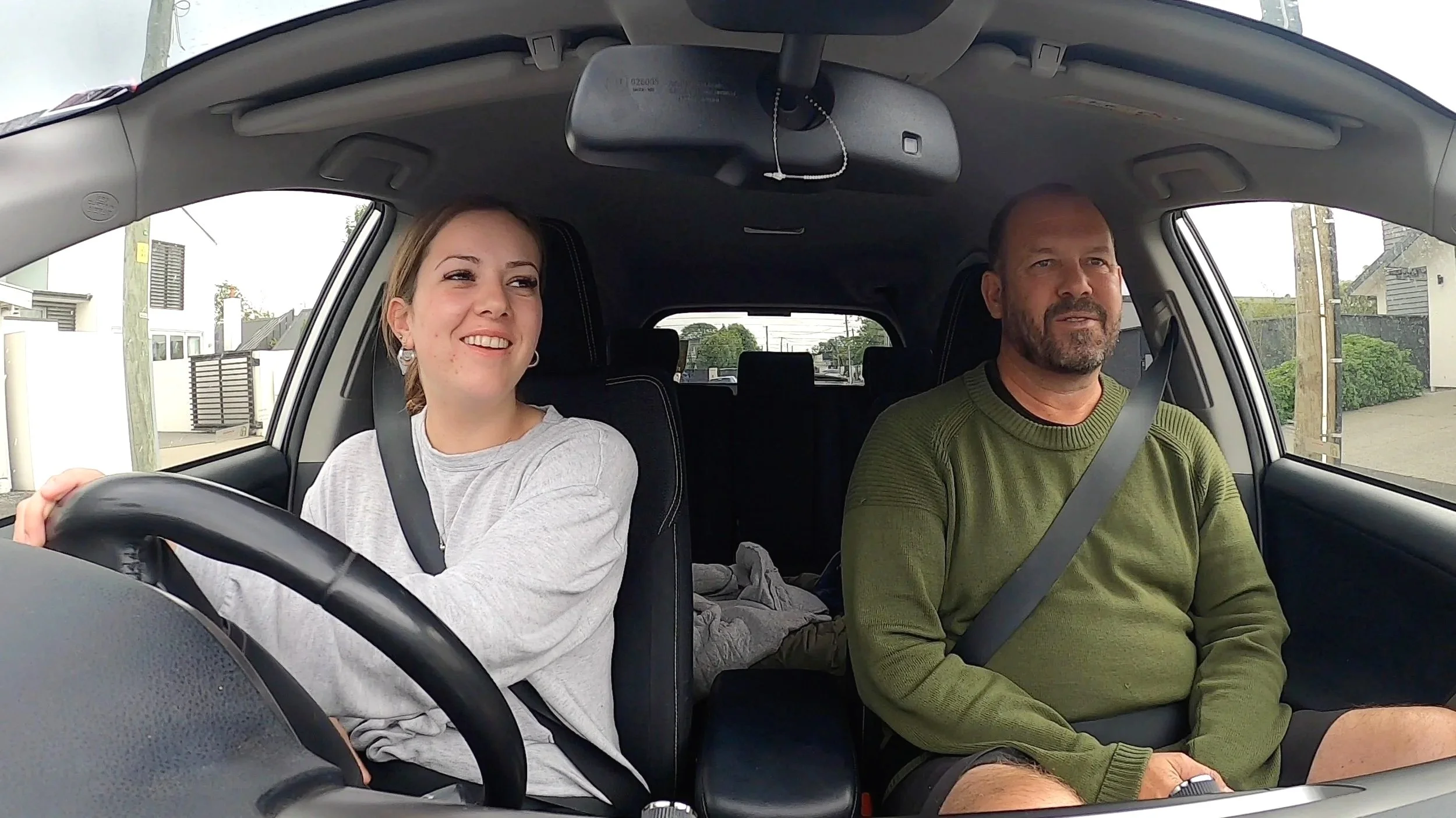 A young woman and older man sitting in a car, with the woman driving and smiling, and the man looking forward, wearing seat belts.