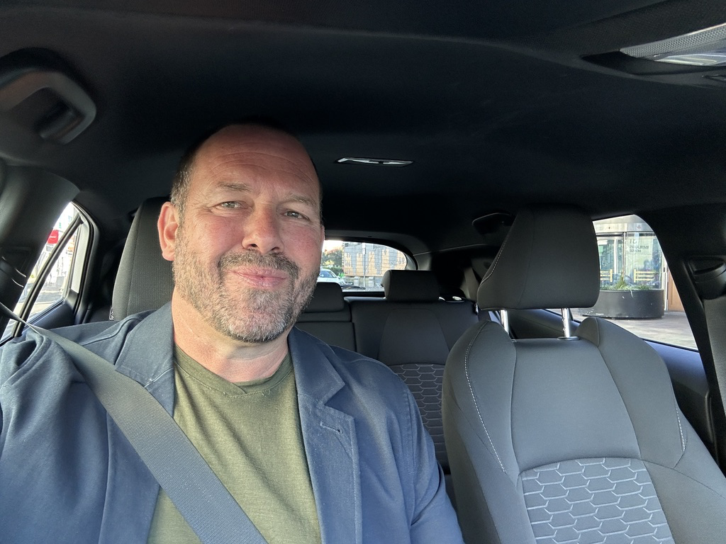 A middle-aged man taking a selfie inside a car, with a seatbelt on, wearing a green shirt and a dark jacket, sitting in the front passenger seat.