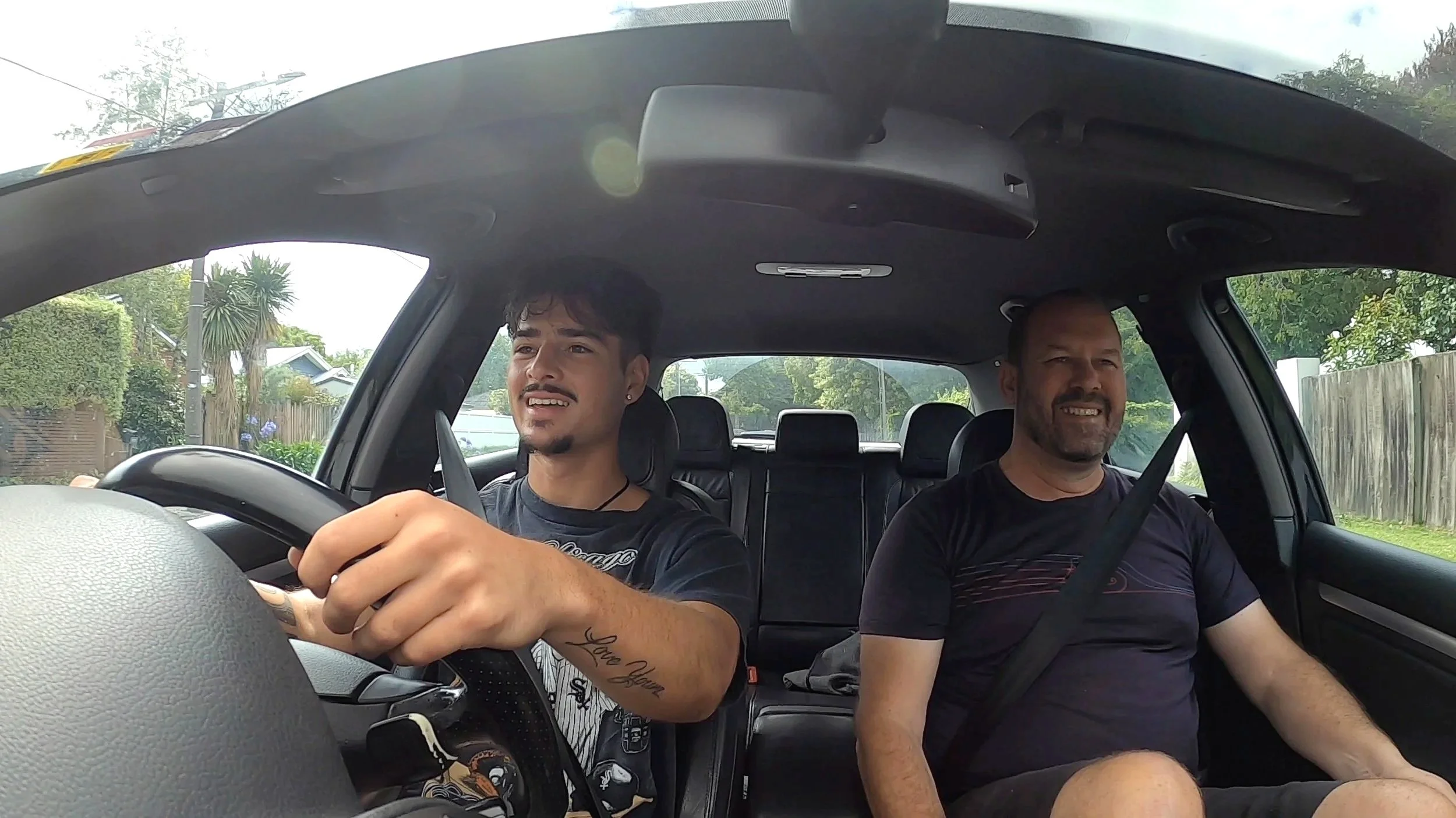 Two men sitting in the front seats of a car, the driver appears to be talking while the passenger is smiling. Outside the windows, greenery and a wooden fence are visible.