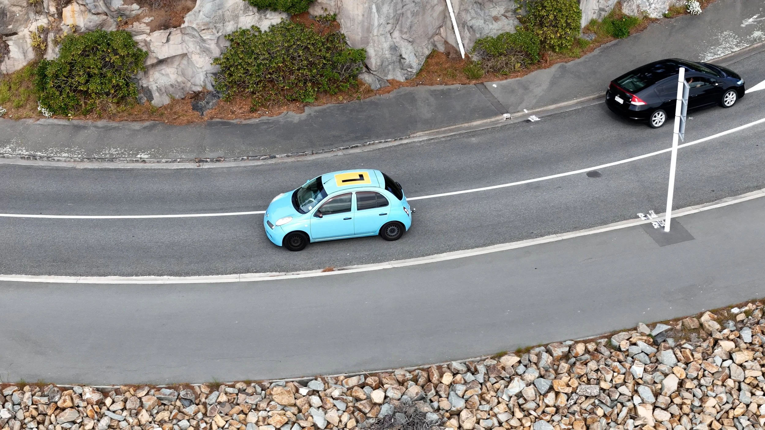 An aerial view of a winding road with two parked cars, a black car and a blue car with a learner's 'L' sign on top, next to a rocky hillside and some greenery.