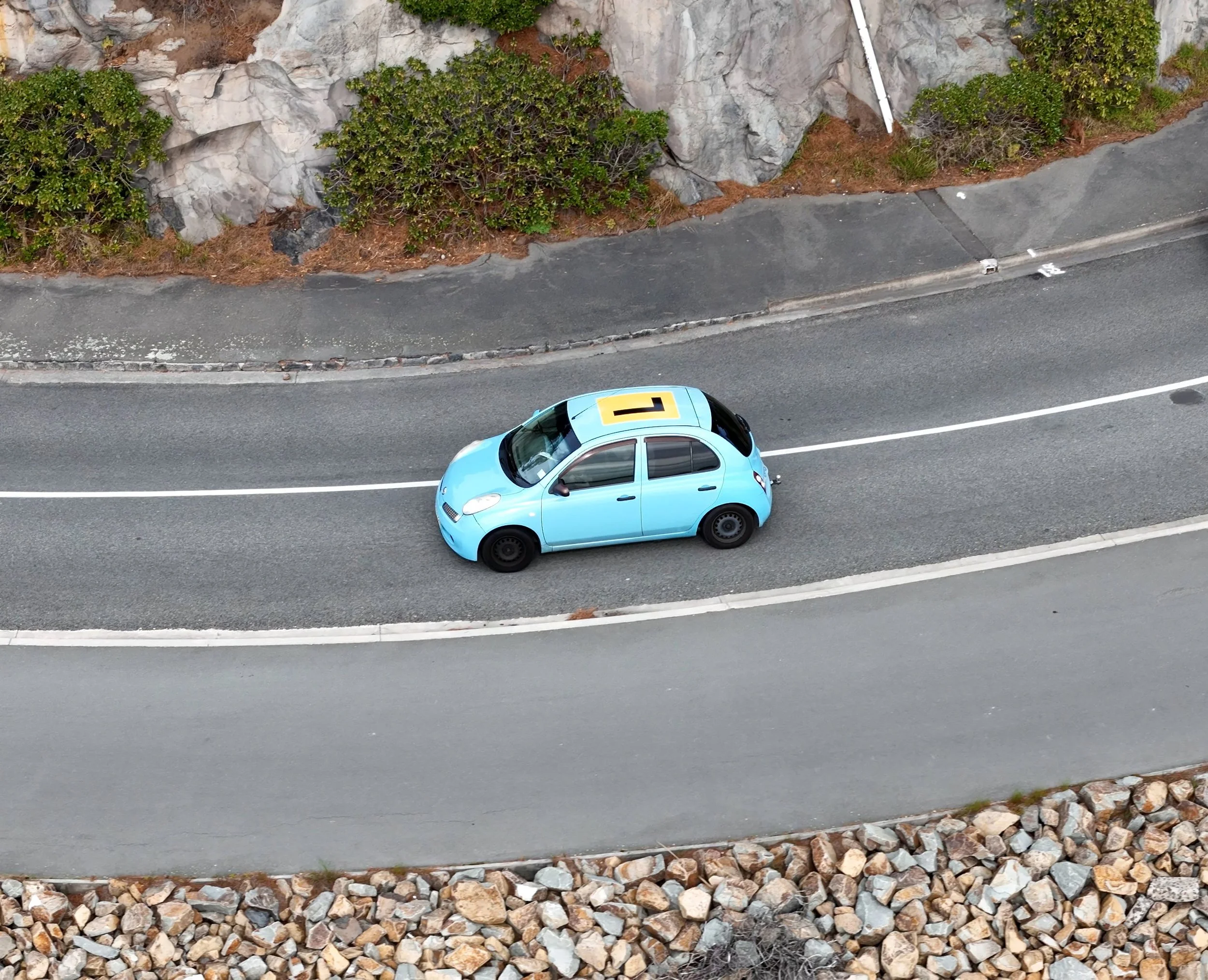 An aerial view of a light blue car on a curved road with rocky and landscaped surroundings.