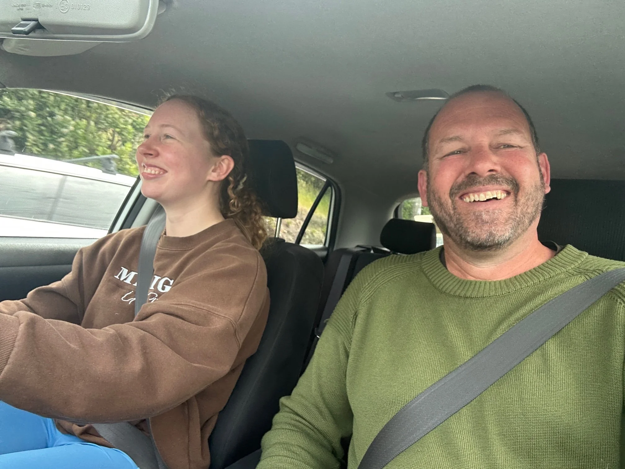 A man and a woman sitting in a car, both wearing seatbelts and smiling happily. The woman is driving and the man is in the passenger seat.