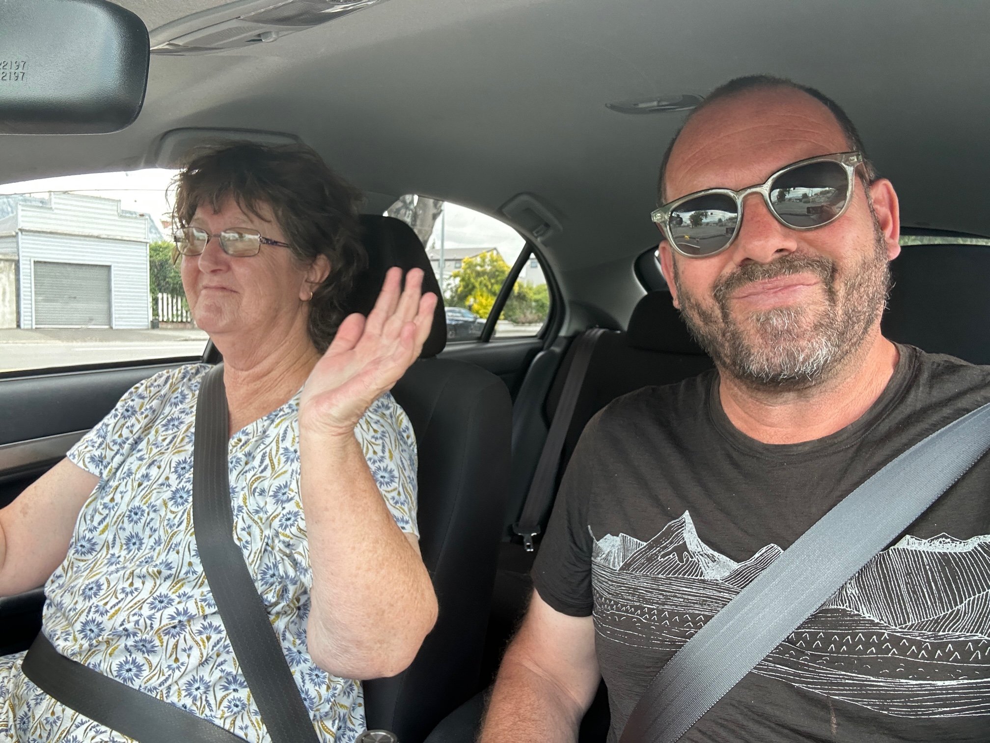 A man and a woman sitting inside a car, both wearing sunglasses. The woman, on the left, is waving and smiling, wearing a floral top, while the man, on the right, has a beard, is smiling, and wearing a black T-shirt with a mountain design. They are both secured with seat belts.