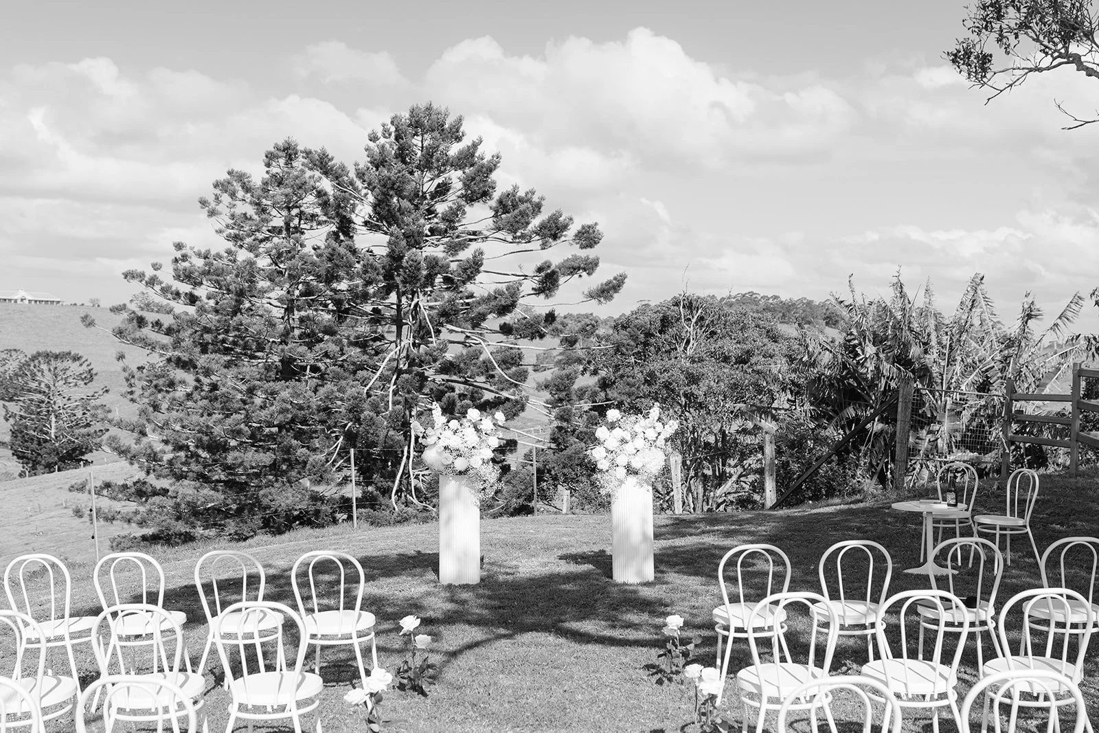 Outdoor wedding setup with chairs arranged in a semicircle and two flower arrangements on tall stands against a backdrop of trees and cloudy sky.