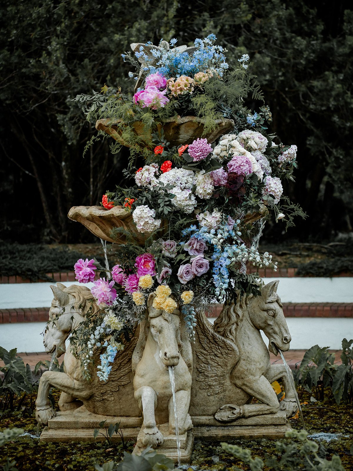 Stone fountain with three mythological horse heads with water flowing from their mouths, adorned with colorful flowers including roses, hydrangeas, and delphiniums, set outdoors with greenery and brick steps in the background.