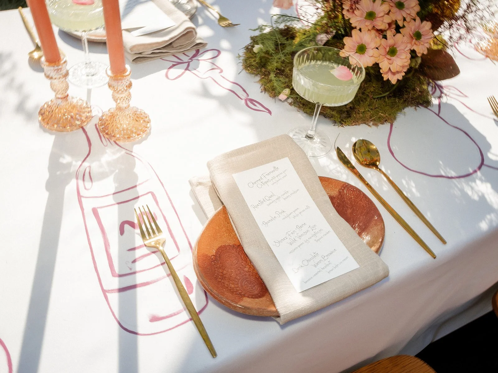 Elegant table setting with gold flatware, pink candlesticks, a menu on a napkin, and a floral centerpiece with pink flowers and greenery.