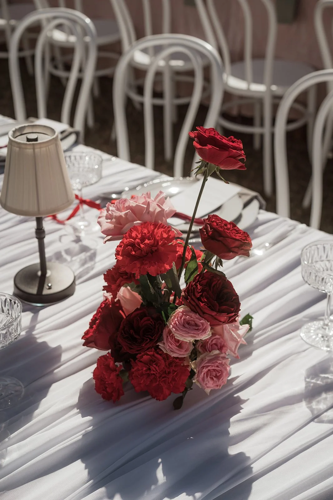 A white tablecloth-covered table decorated with a pink and red floral centerpiece, clear glassware, and a small table lamp, set outdoors with white chairs in the background.
