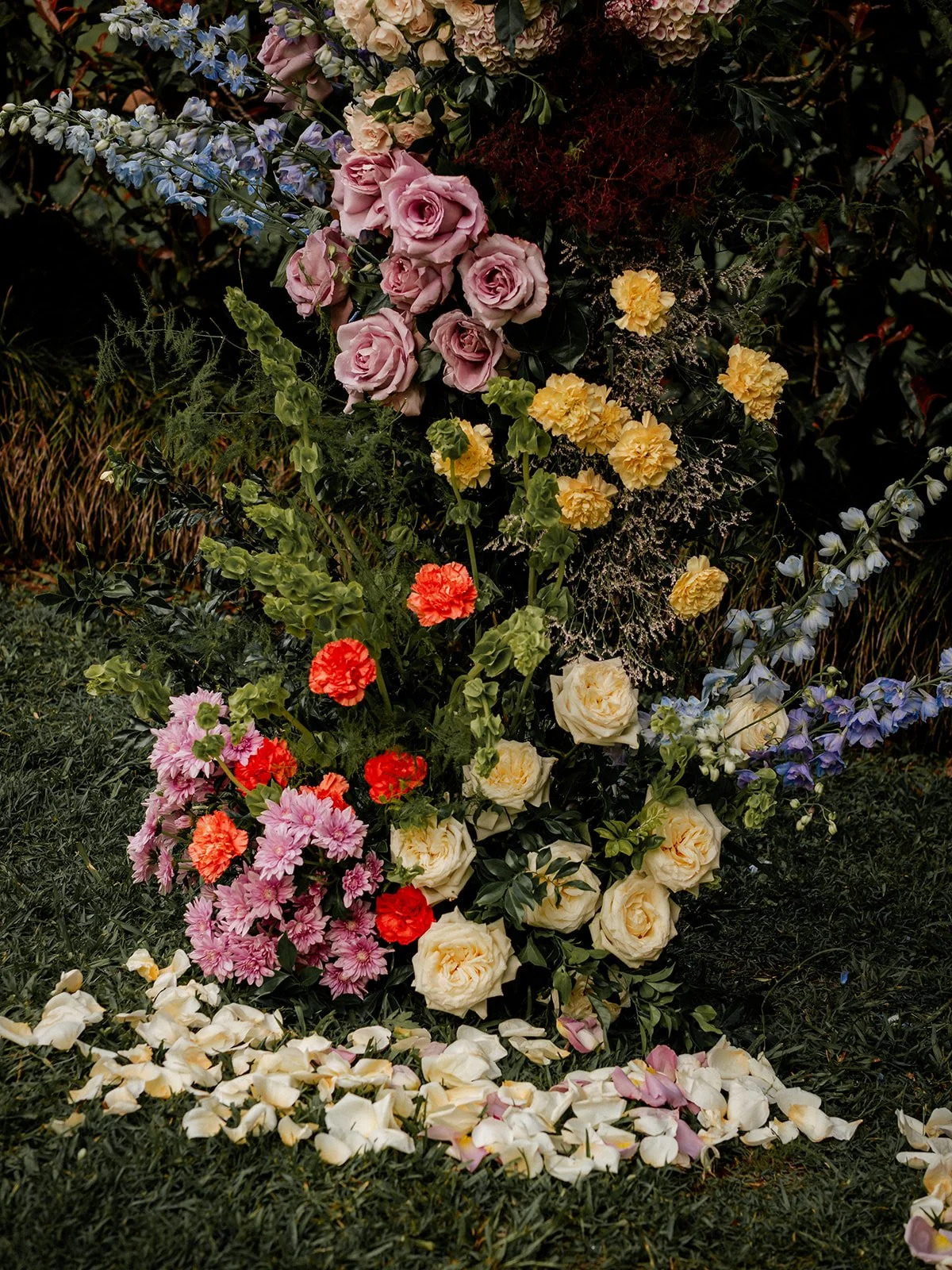 A floral arrangement with pink, yellow, white, red, and purple flowers on green grass with scattered rose petals at the bottom.