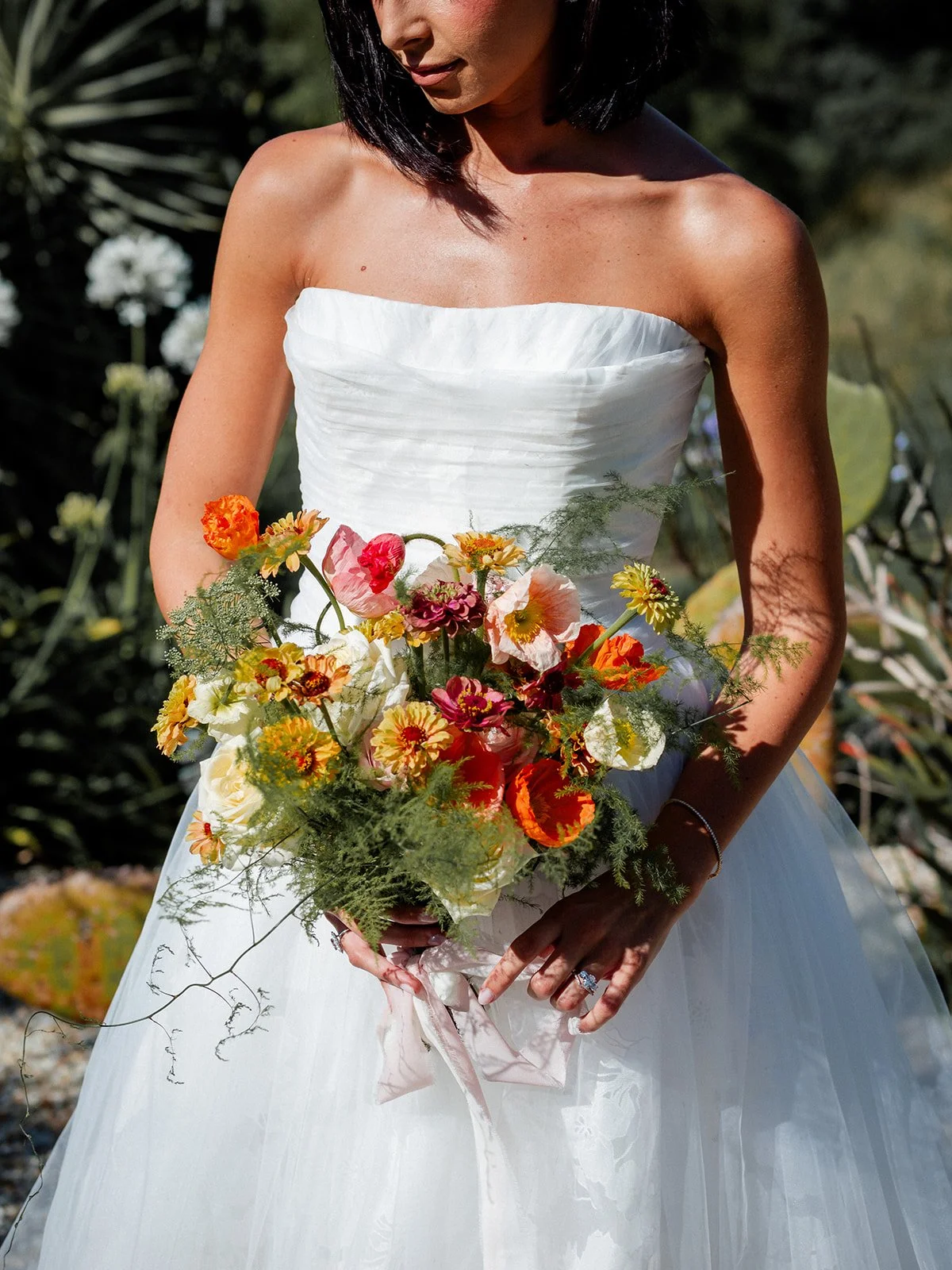 A woman in a strapless white wedding dress holding a colorful bouquet of flowers outdoors.
