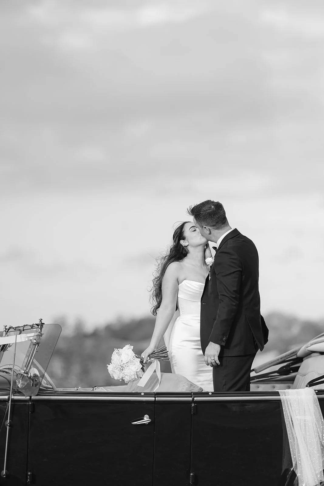 A black and white photo of a bride and groom sharing a kiss in a vintage car, with she waved veil and a bouquet of flowers, outdoors on a cloudy day.