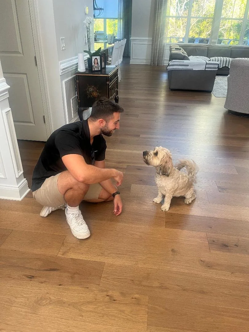 A man crouching and smiling at a small, curly-haired dog indoors in a living room.