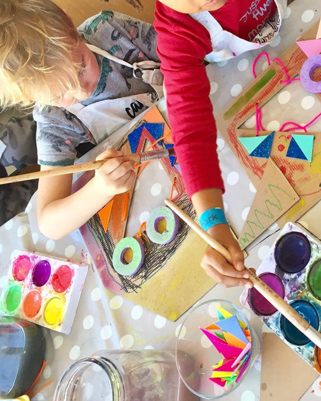 Children engaged in arts and crafts, painting, and decorating paper with bright colors, tape, and foam rings on a polka-dot tablecloth.