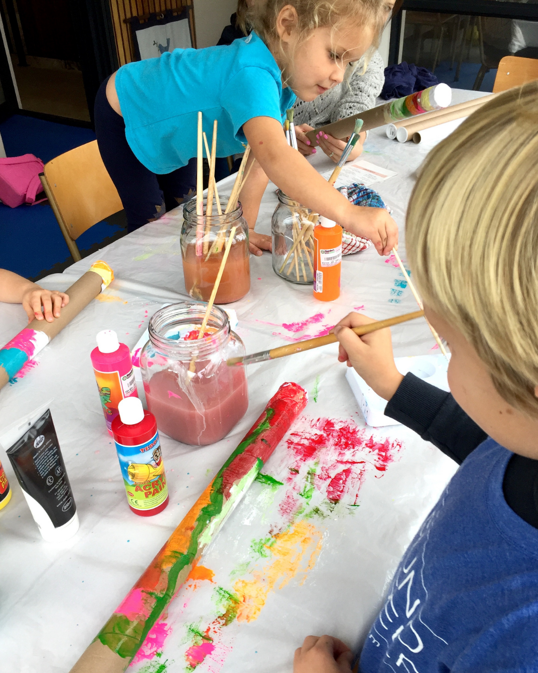 Children craft with painted cardboard tubes and paint, using brushes and glue at a table covered with a white sheet.