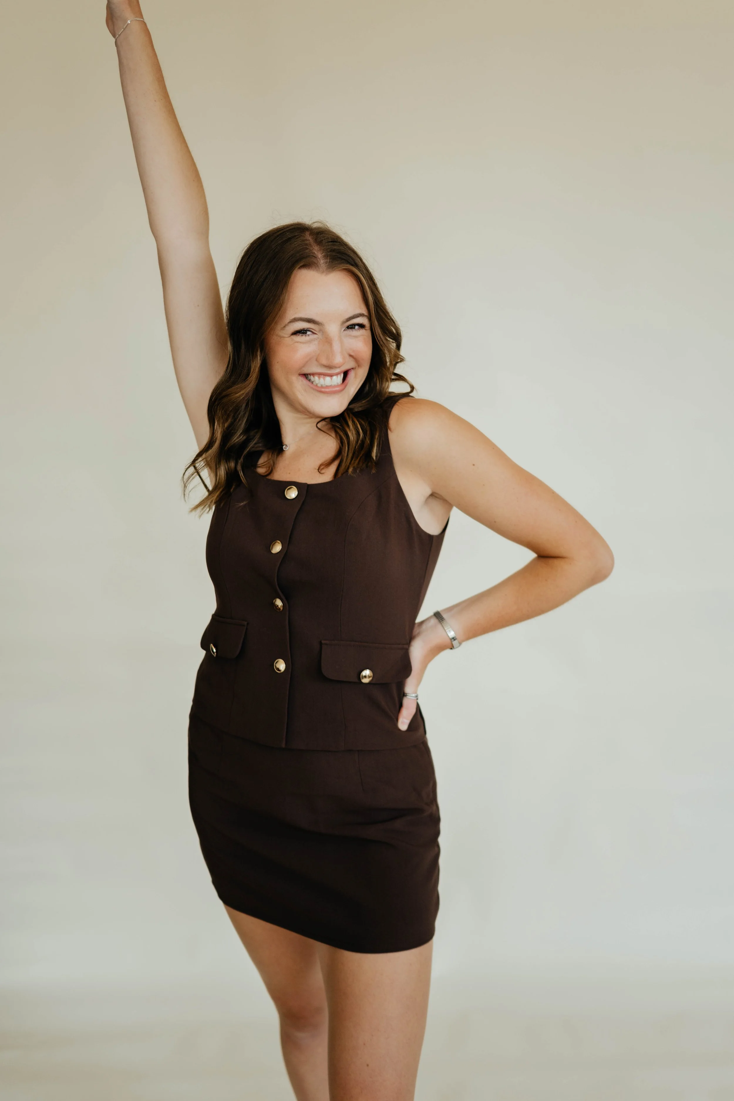 A young woman with brown hair wearing a sleeveless brown dress with gold buttons, smiling, standing against a plain light-colored background.