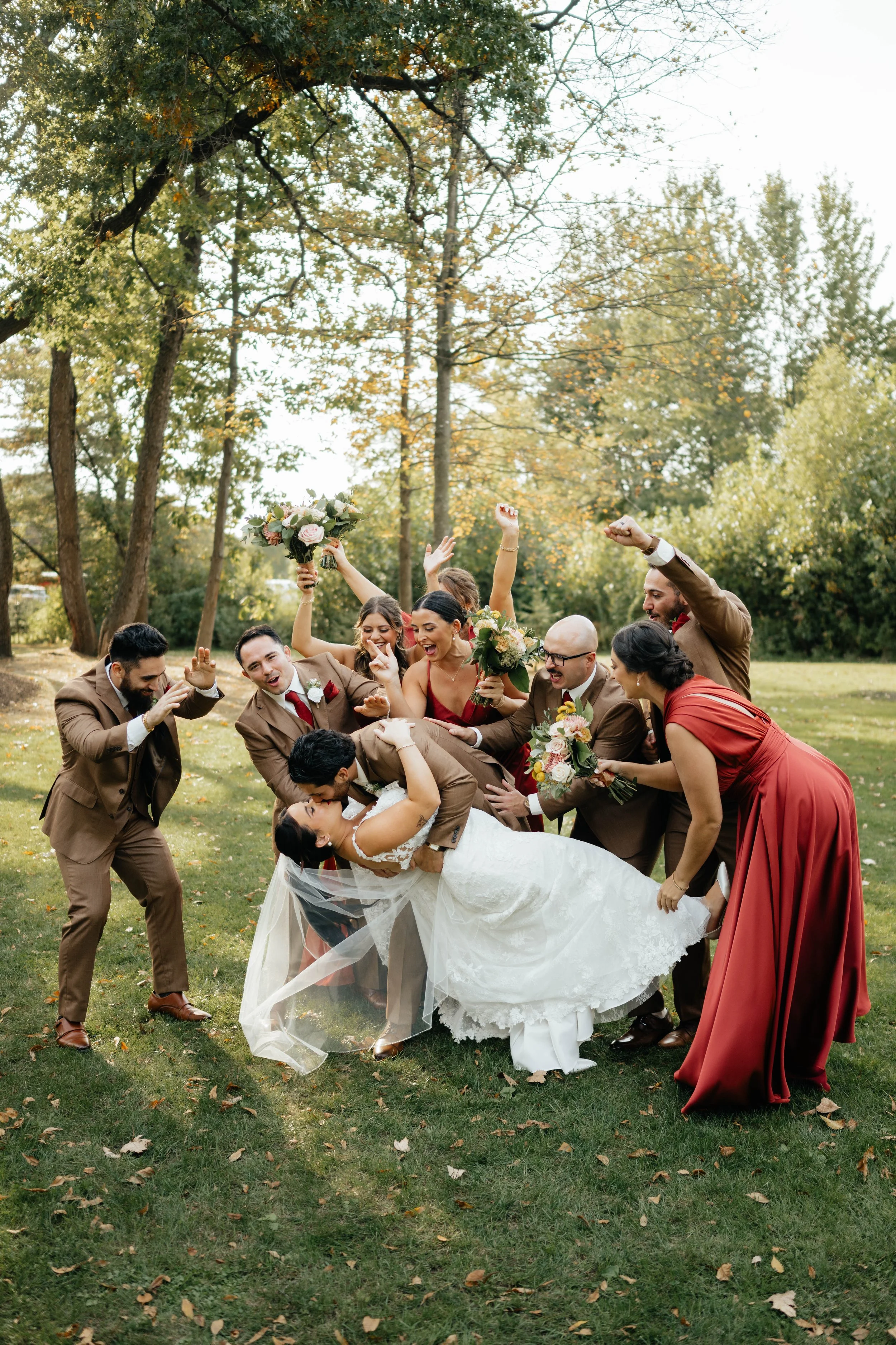 A bride and groom are surrounded by wedding guests outdoors, with the bride being dipped slightly and kissed while everyone around cheerfully celebrates and holds flowers, in a park with trees.
