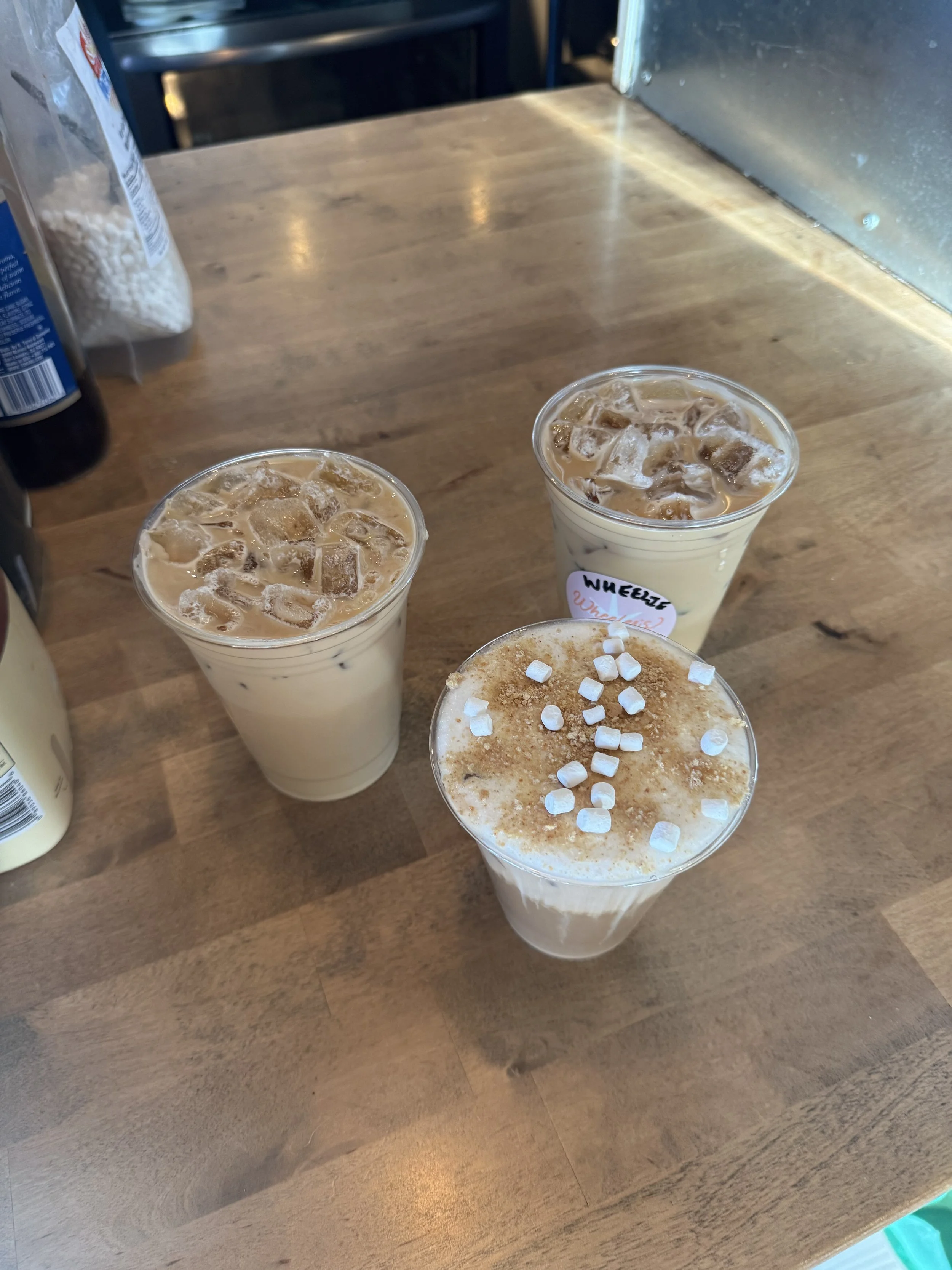 Three iced beverages with toppings on a wooden table, with a plastic bottle, bag of marshmallows, and soda bottle in the background.