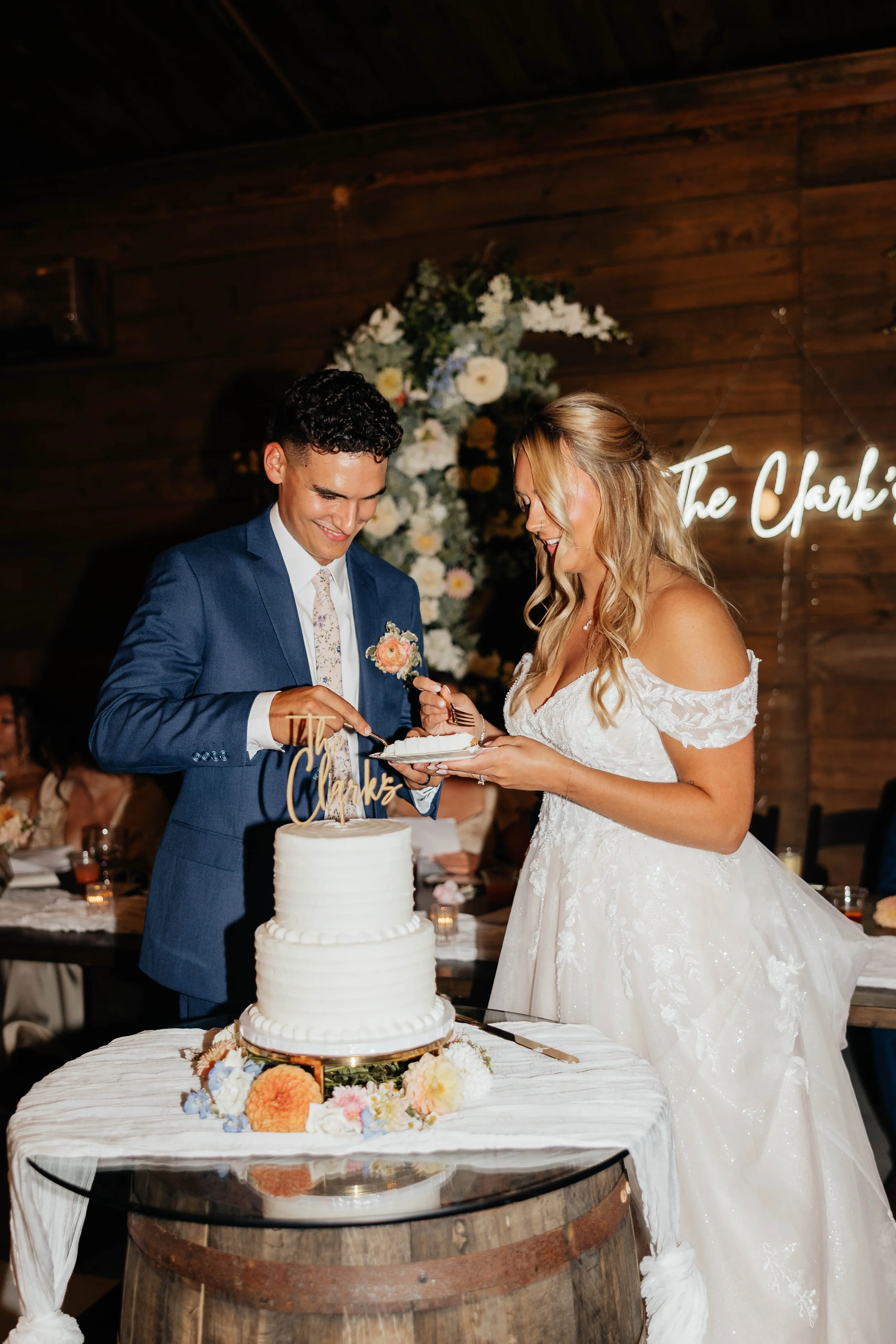 A newlywed couple is cutting a wedding cake in a rustic venue, with a floral arch and neon sign in the background.