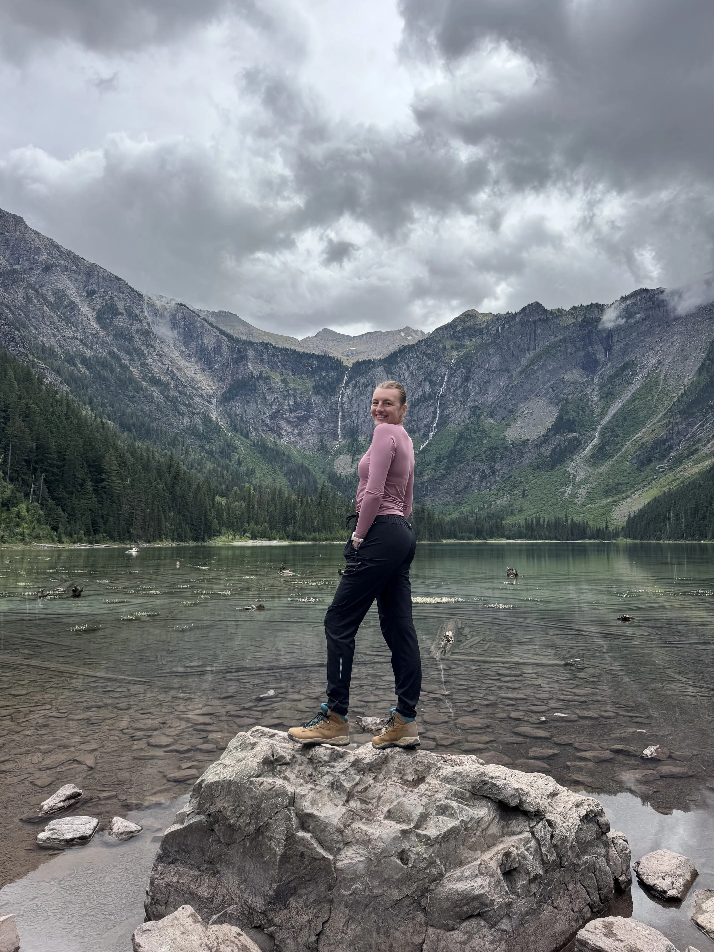 A woman standing on a large rock in a lake with mountains and waterfalls in the background under cloudy skies.