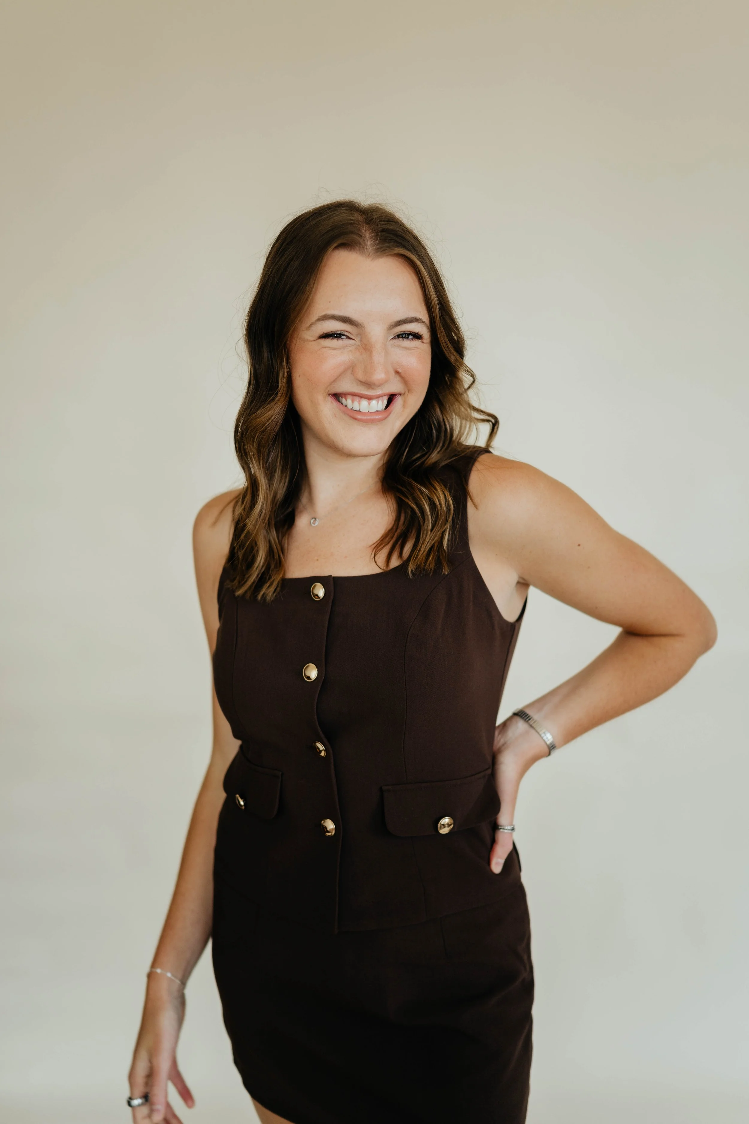 A smiling woman with wavy brown hair wearing a sleeveless black dress with gold buttons, standing against a plain beige background.