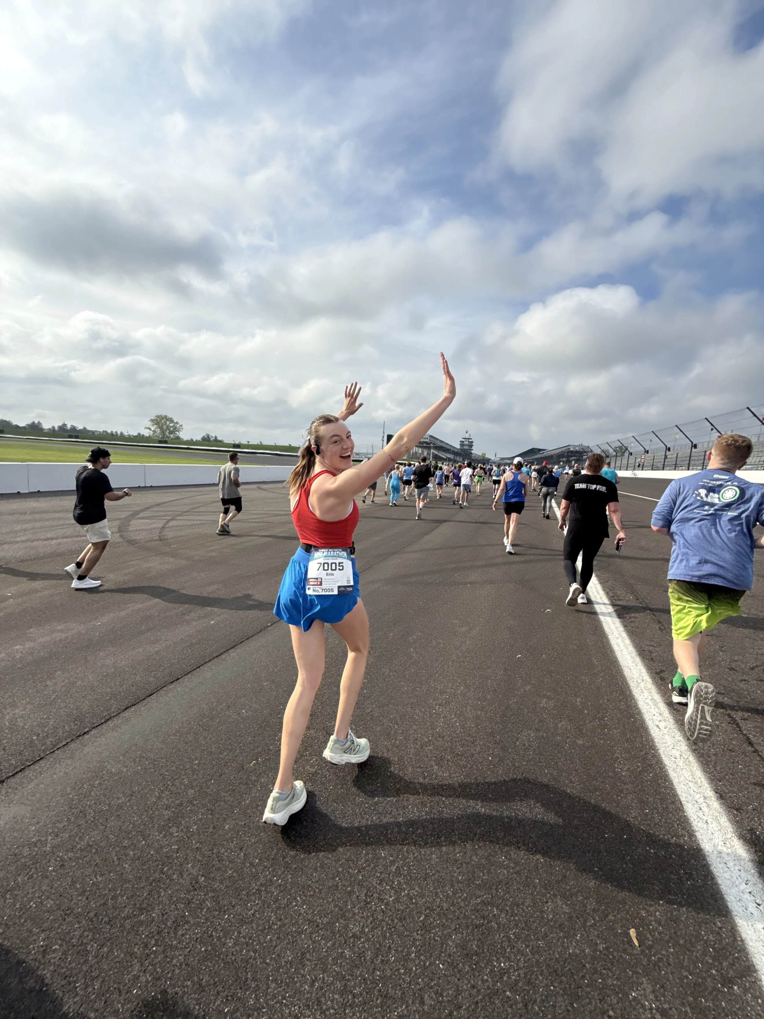 A woman in a red tank top and blue shorts smiling and waving at a marathon race, with other runners on a race track under a partly cloudy sky.