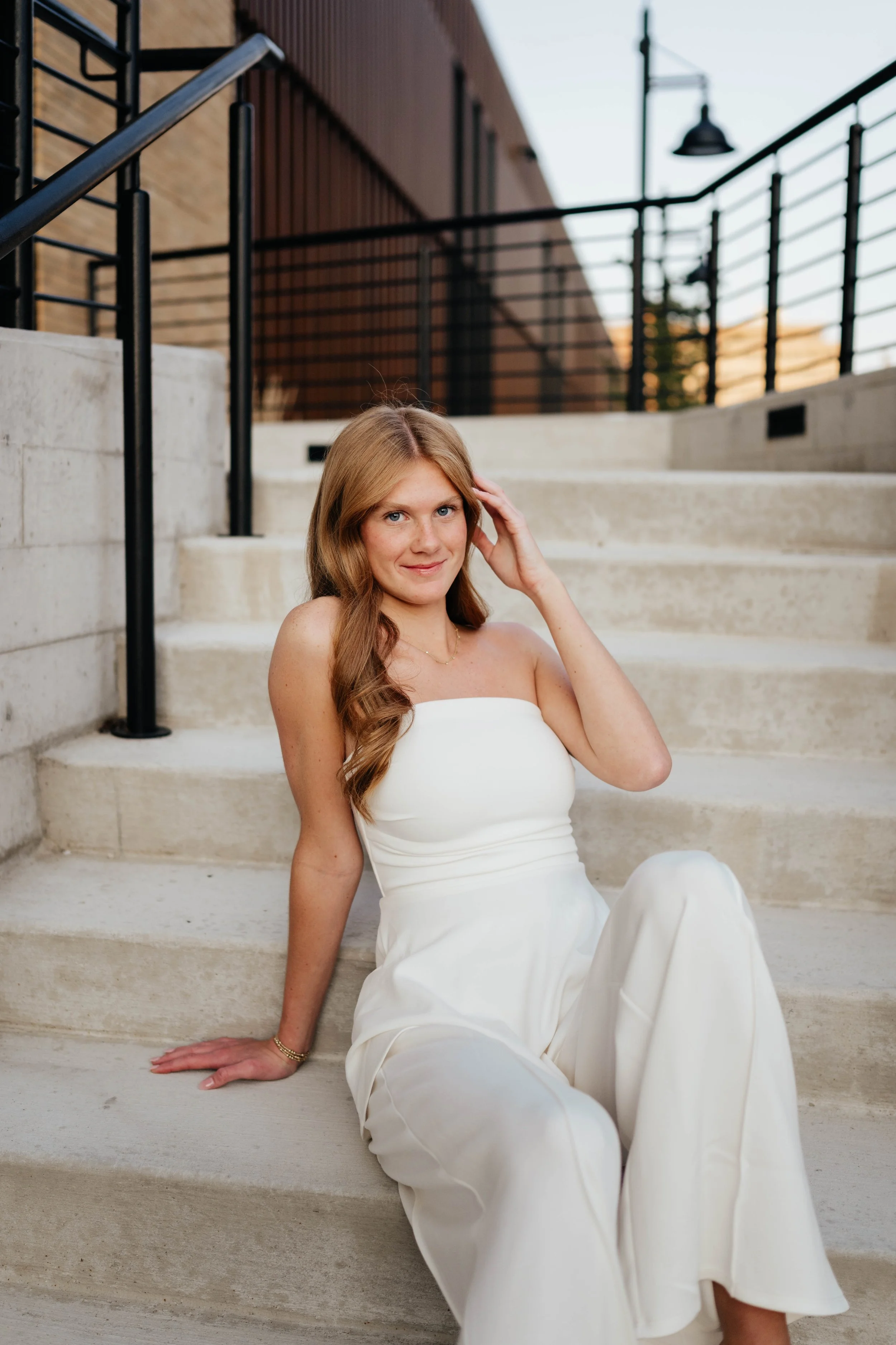 A woman in a white strapless jumpsuit sitting on concrete stairs outdoors, touching her hair and smiling at the camera.