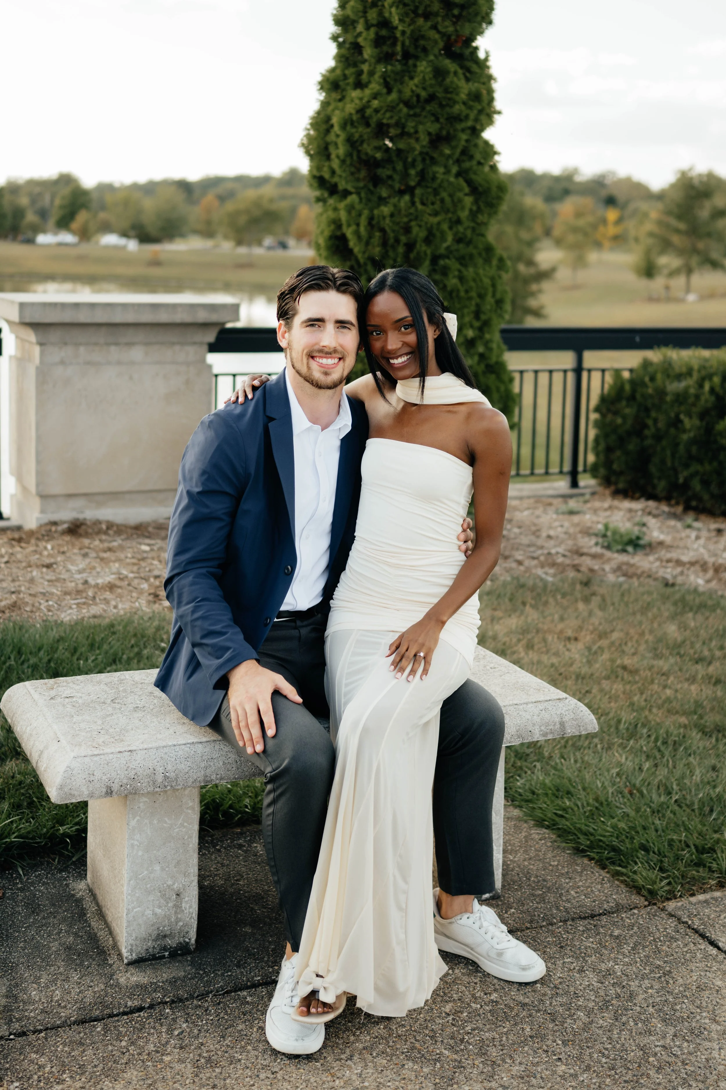 A smiling couple sitting on a concrete bench outdoors, with the woman in a white dress and the man in a blue blazer and white shirt, surrounded by greenery and trees.