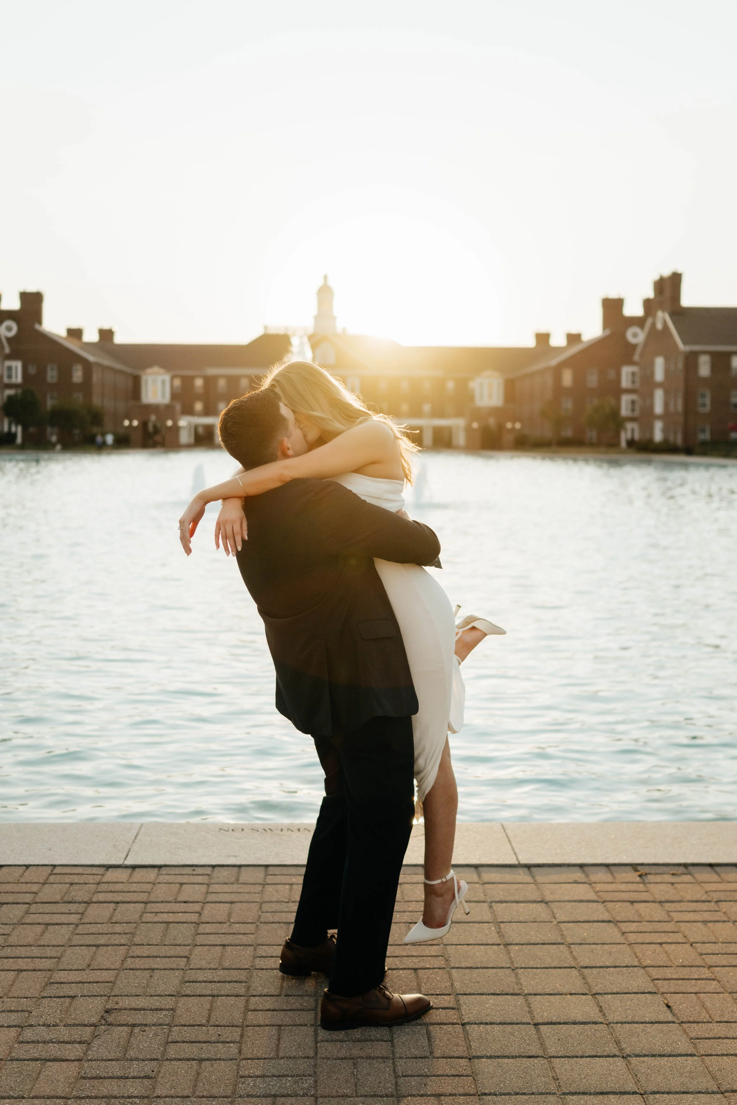 A man lifting a woman in a white dress by a lake at sunset, with buildings in the background.