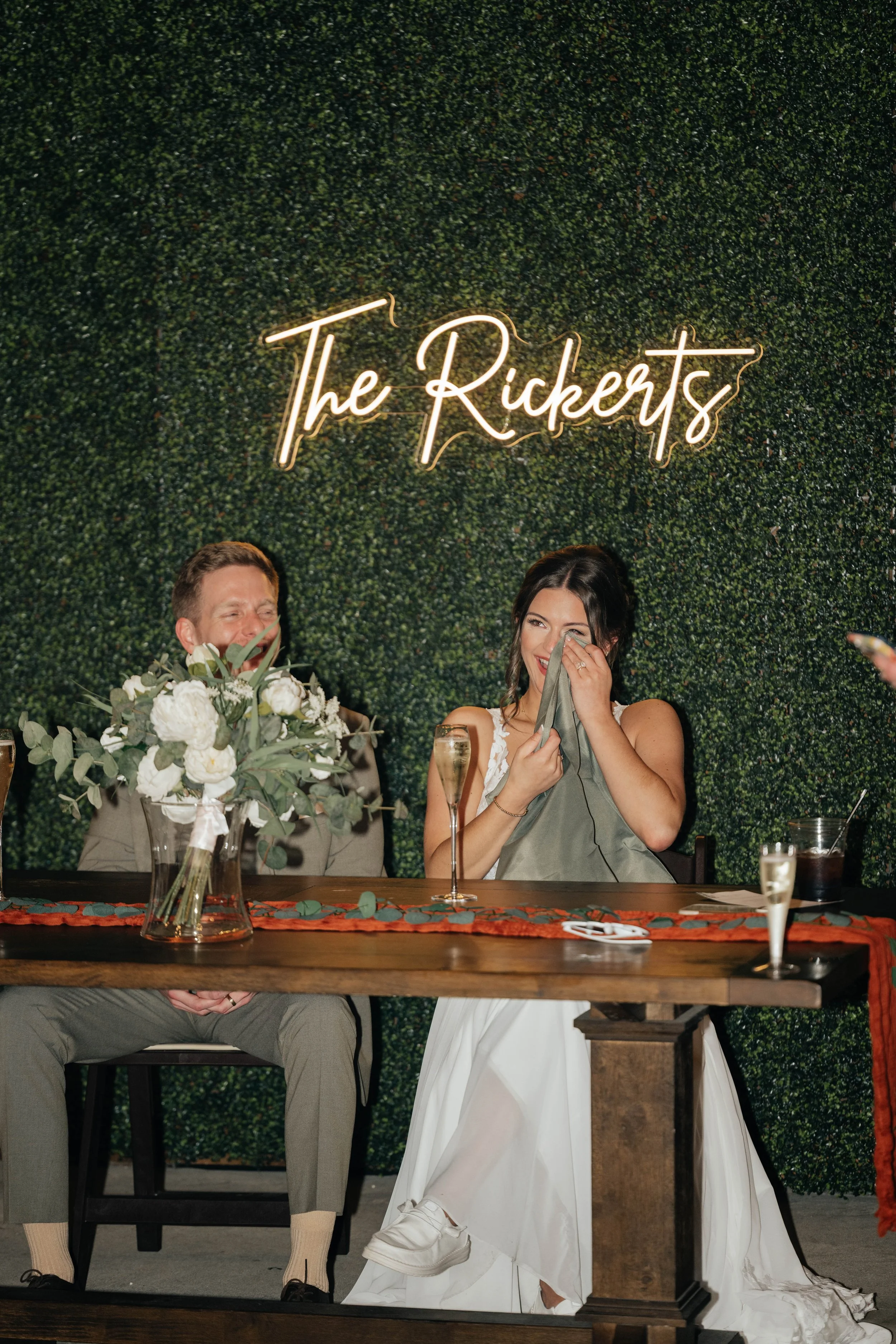 A bride and groom sitting at a decorated table during their wedding reception, with a green hedge wall backdrop featuring a neon sign that reads 'The Ricketts'. The bride is laughing, holding a cloth to her face, and the groom is smiling. The table has a bouquet of white flowers and glasses of champagne.