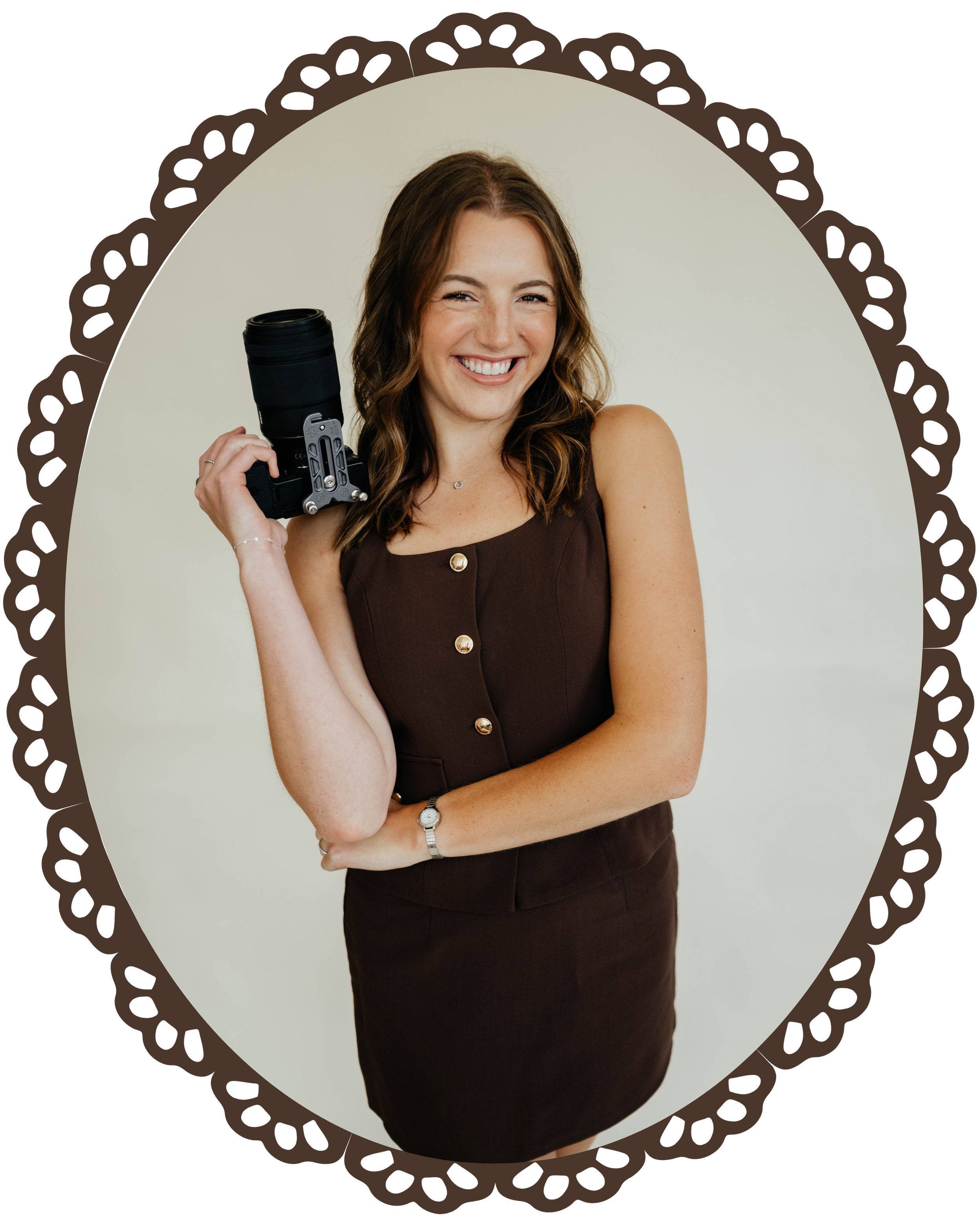 A smiling woman holding a camera with a large lens, wearing a sleeveless brown dress with gold buttons, posing against a plain light background.