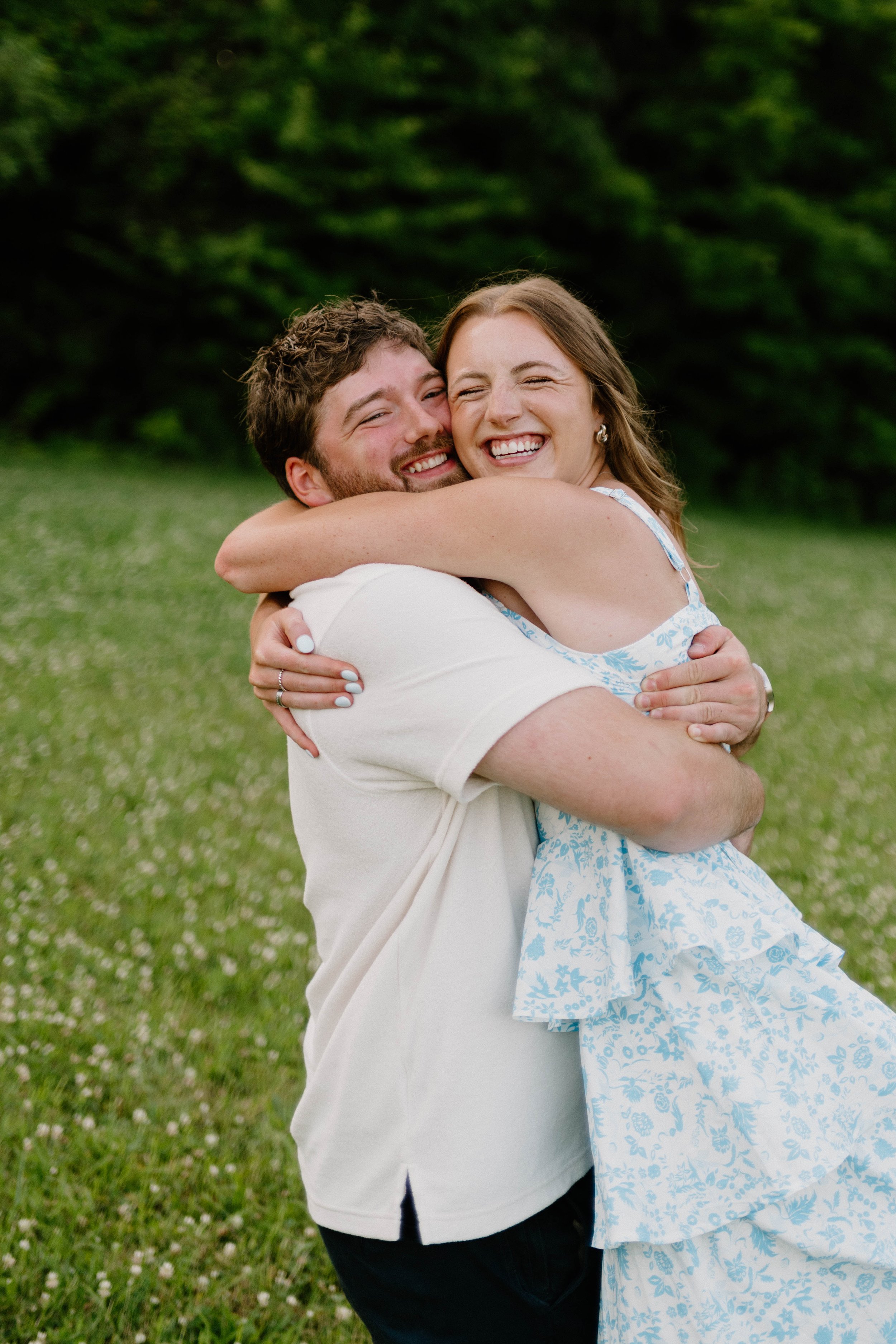 A happy couple hugging outdoors in a grassy field, smiling and laughing with trees in the background.