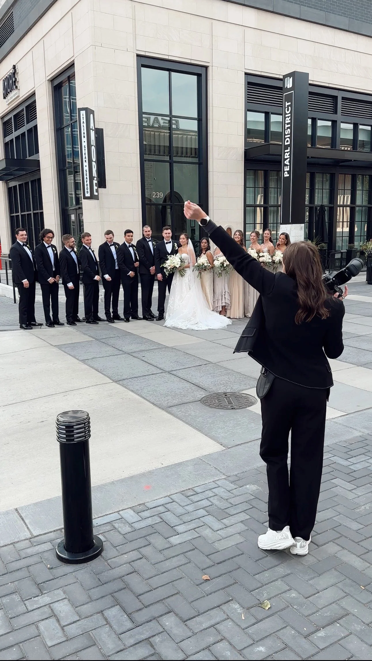 A group of wedding party members, including the bride, groom, and bridesmaids, pose for a photo outdoors in front of a modern building with large windows and signs that read 'Pearl District' and 'Il Forno.' An individual with a camera is taking their picture, standing on a brick sidewalk.