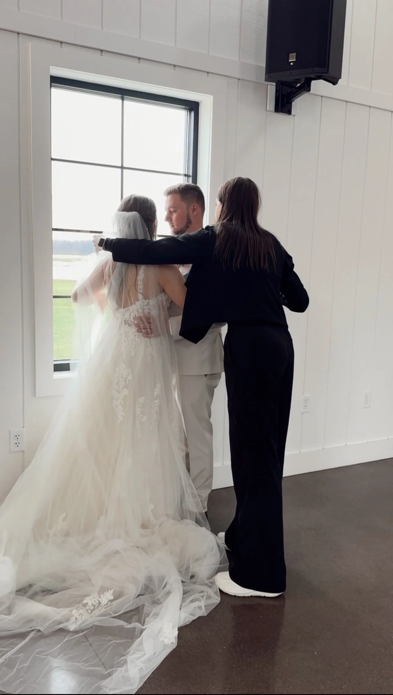 Bride and groom embracing near a window while a woman in black adjusts the bride's veil during a wedding ceremony in a bright, white-walled room.