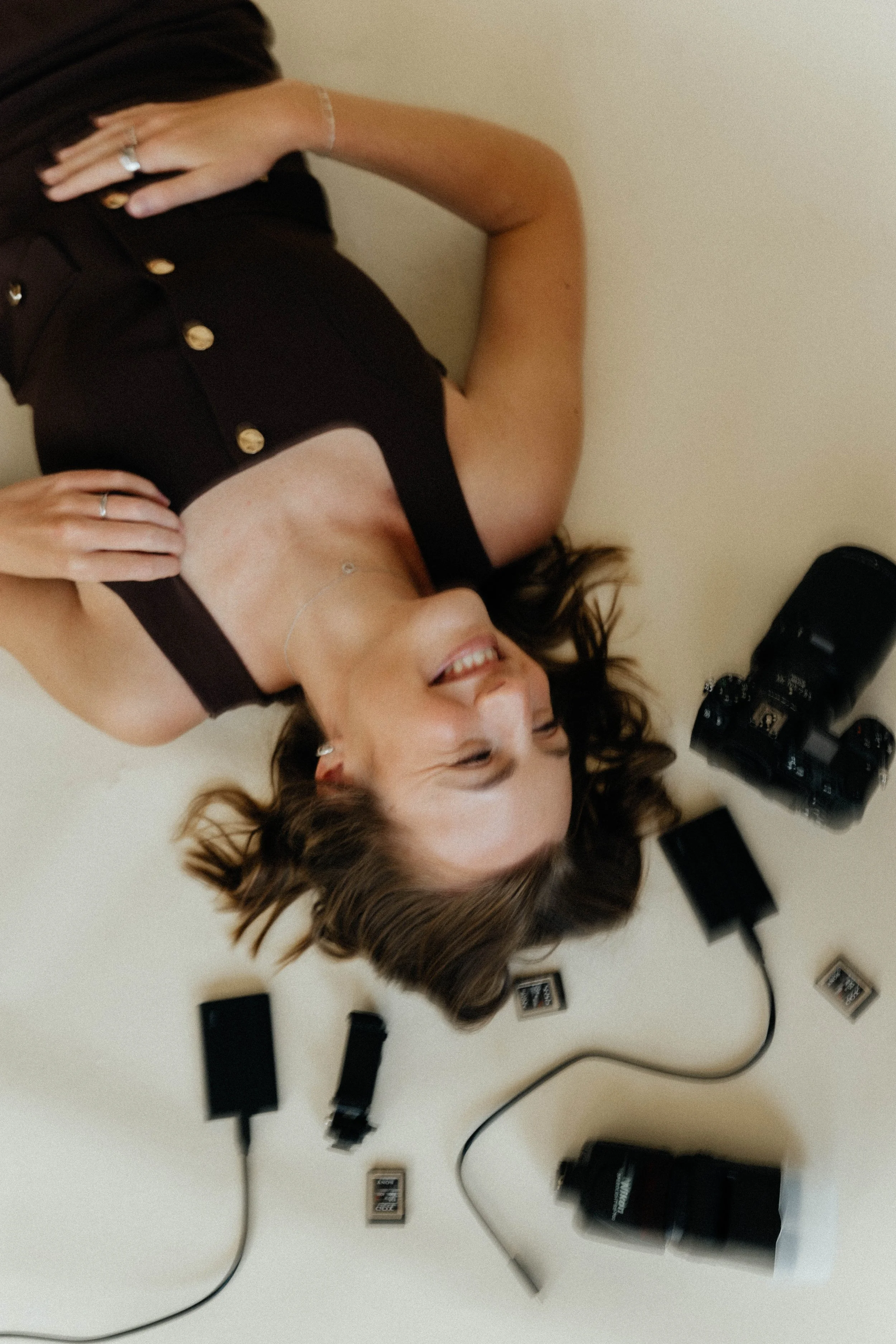 Woman lying on the floor, surrounded by camera equipment and memory cards, smiling and looking upward, wearing a black dress.
