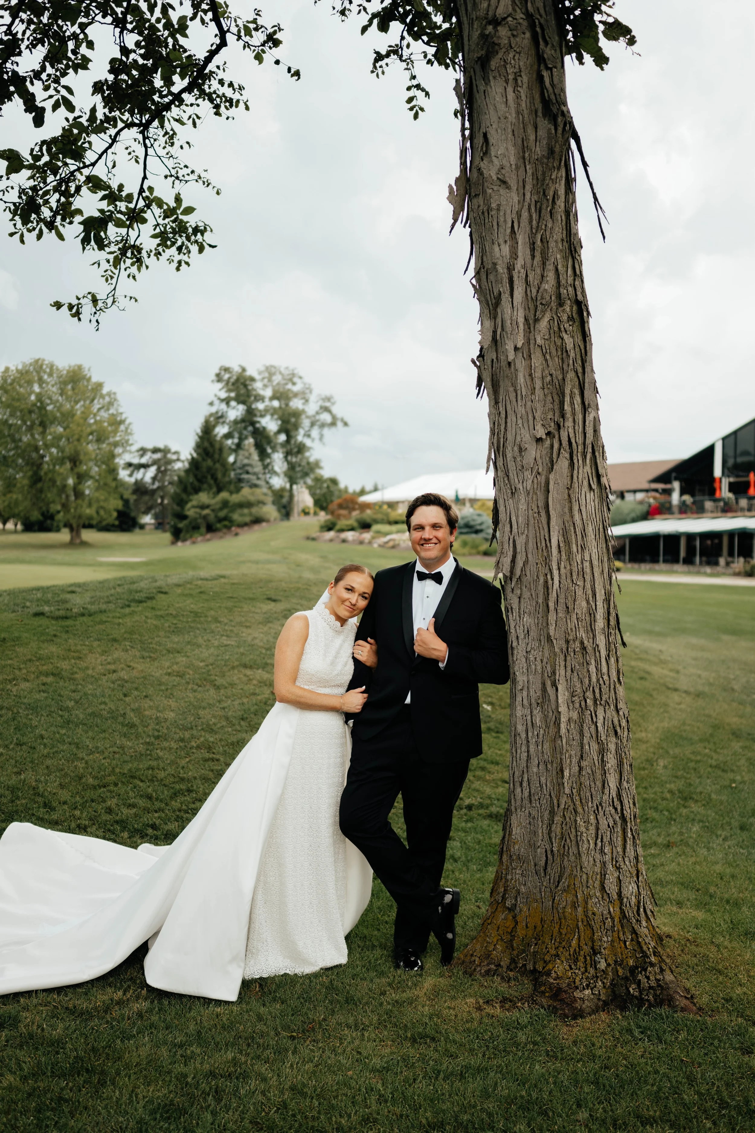A bride and groom in wedding attire standing outdoors near a large tree, smiling at the camera with a backdrop of greenery and a building.